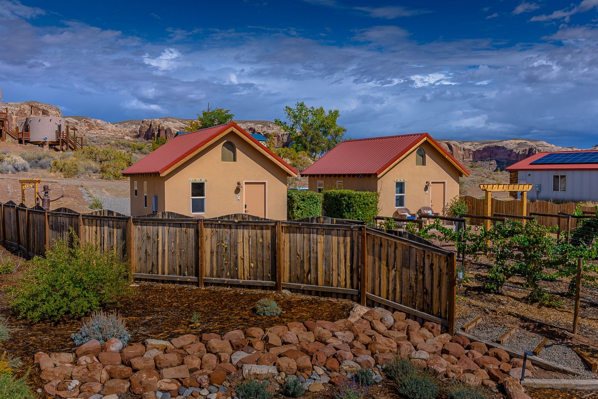 Two tan cottages with red roofs behind a wooden fence, nestled in a rocky landscape under a cloudy sky.