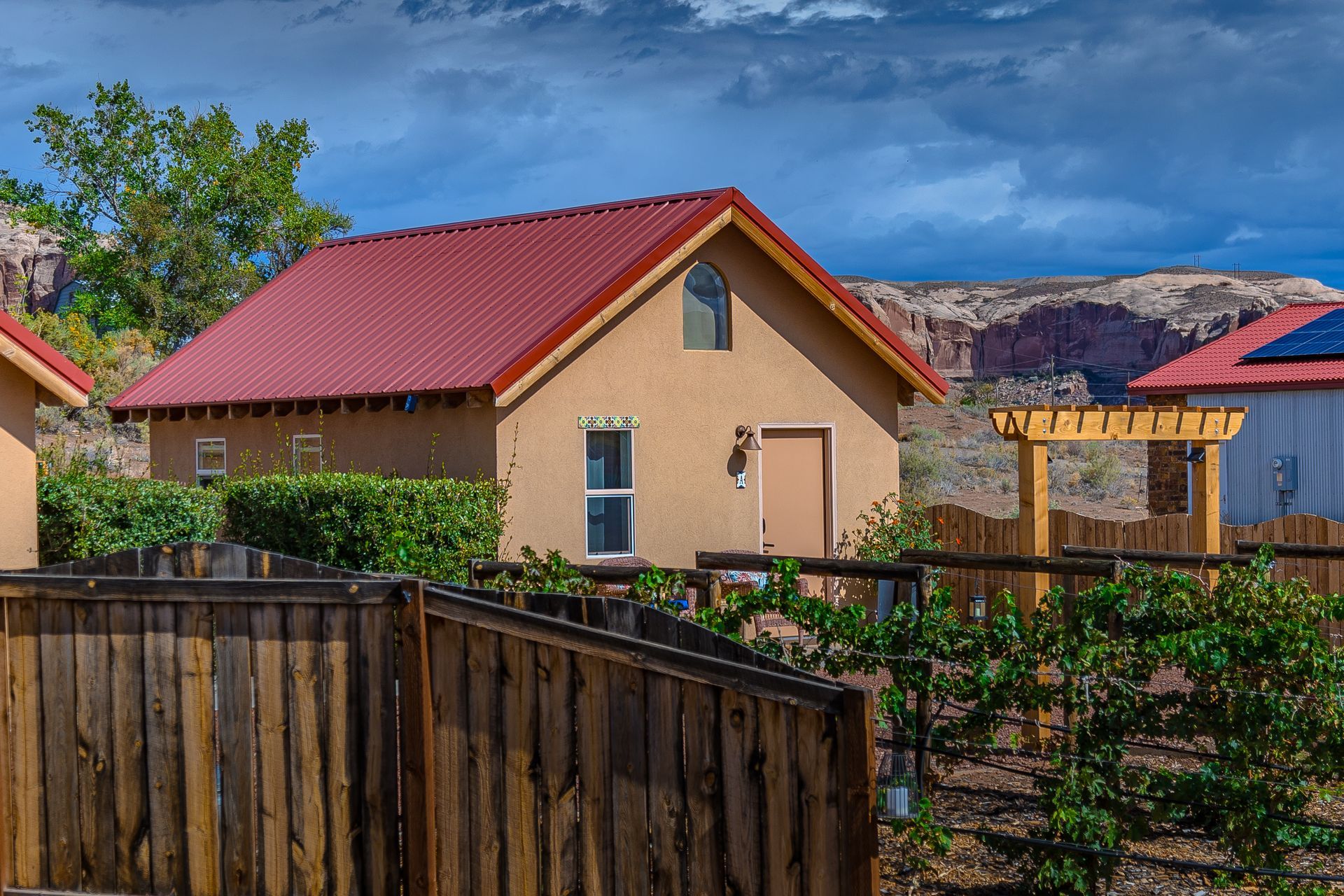 Tan adobe cottage with a red metal roof and wooden fence, set against a desert landscape.