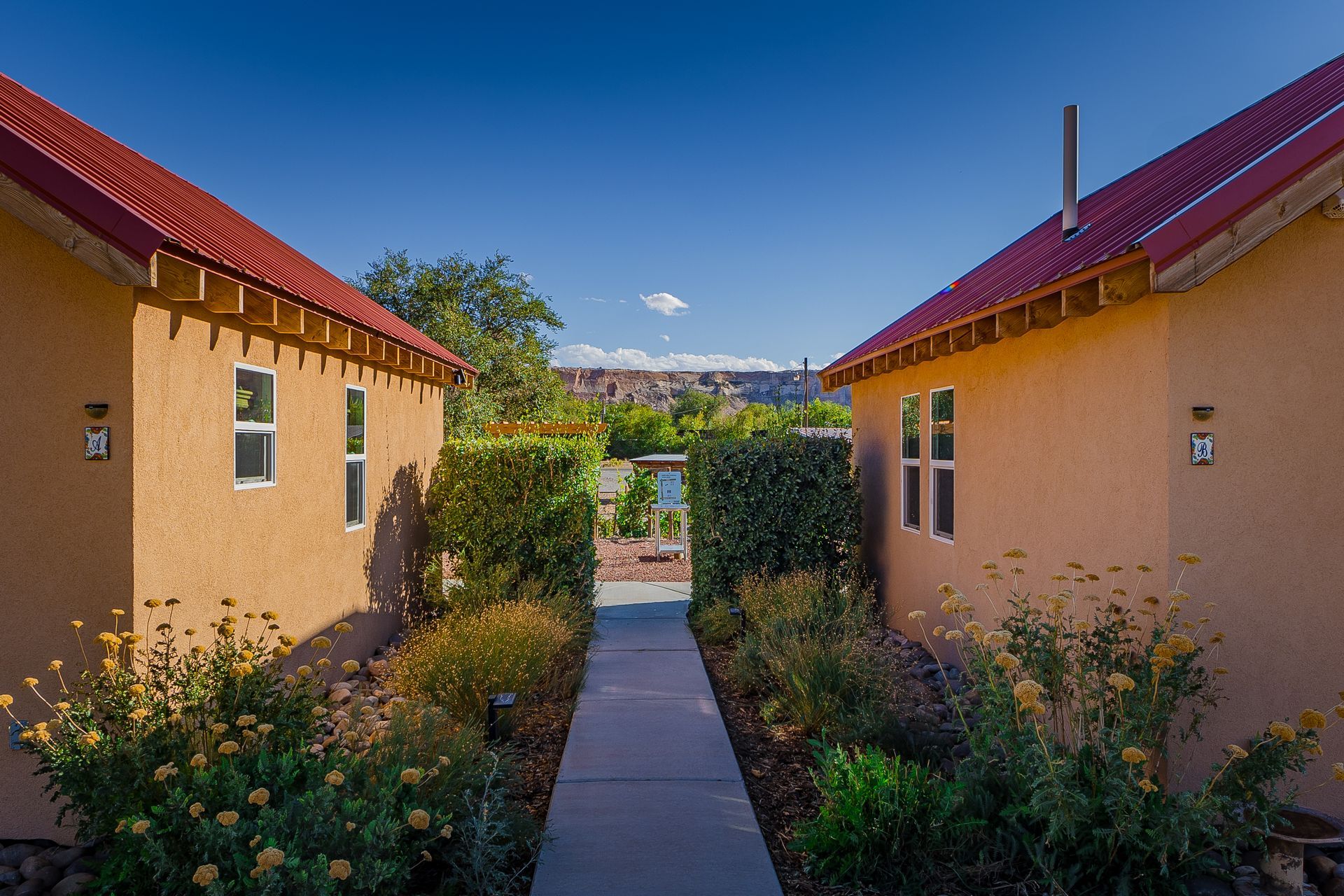 Two tan buildings with red roofs frame a walkway lined with bushes and flowers, under a blue sky.