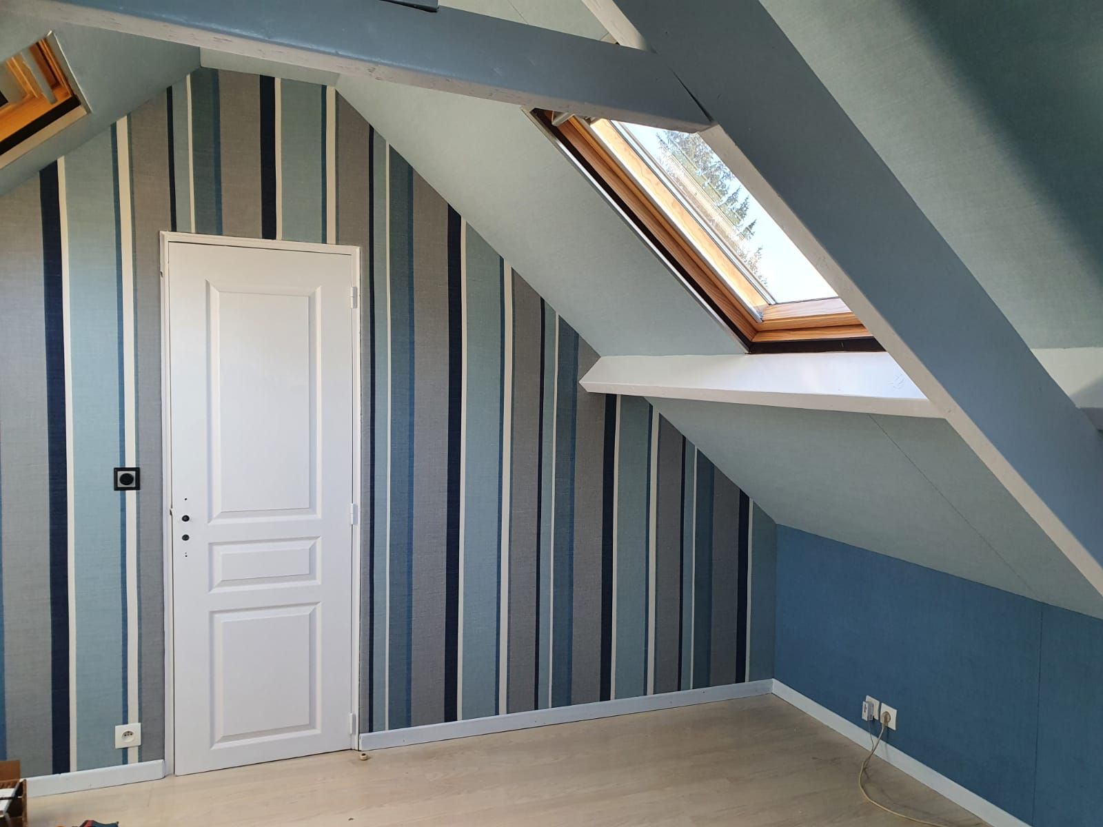 Chambre mansardée avec papier peint à rayures dans les tons bleu et gris. Porte blanche et plafond mansardé avec puits de lumière.