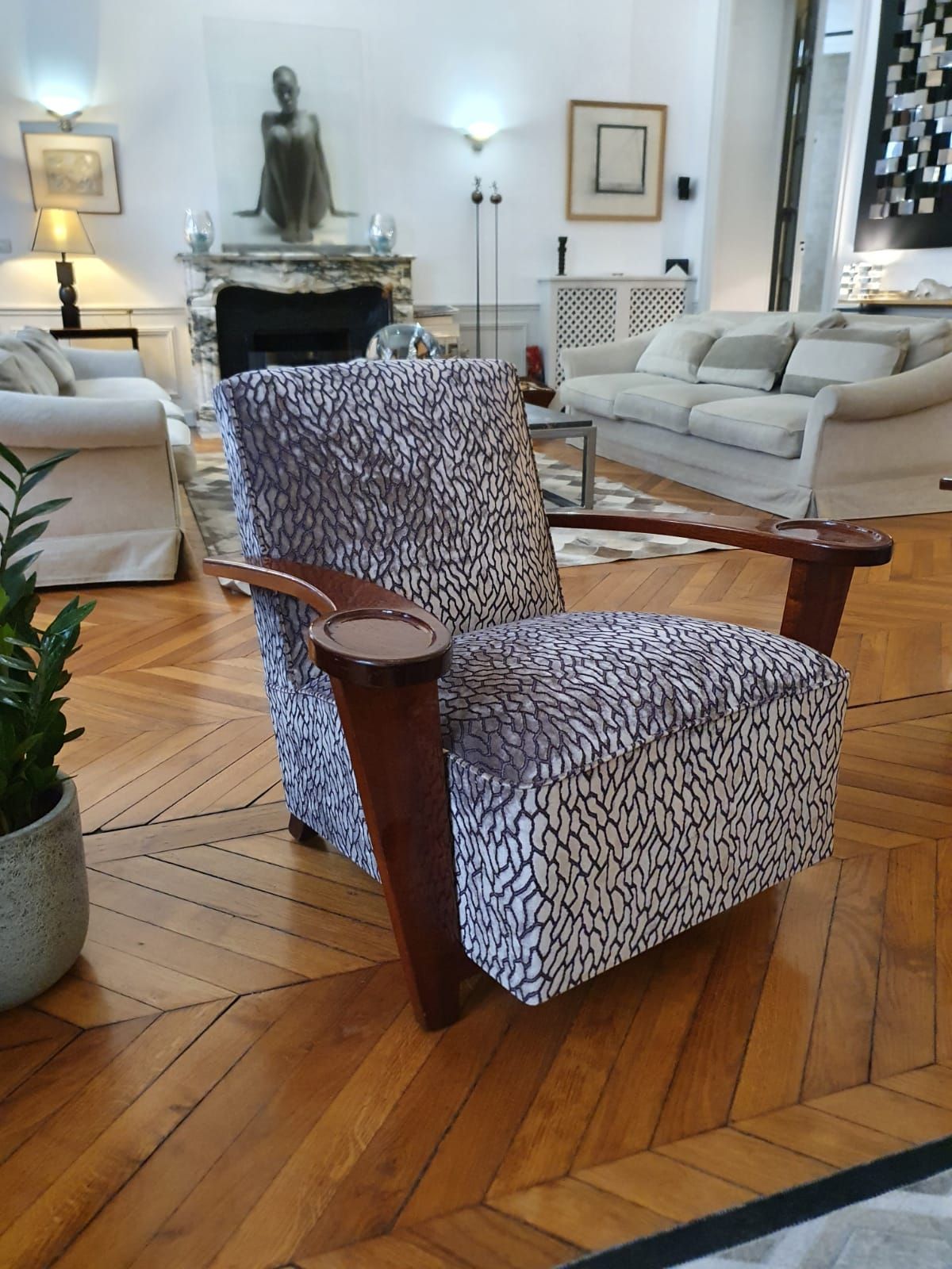 Un fauteuil à motifs avec accoudoirs en bois repose sur un parquet. À l'arrière-plan, un salon avec des canapés blancs et une cheminée.