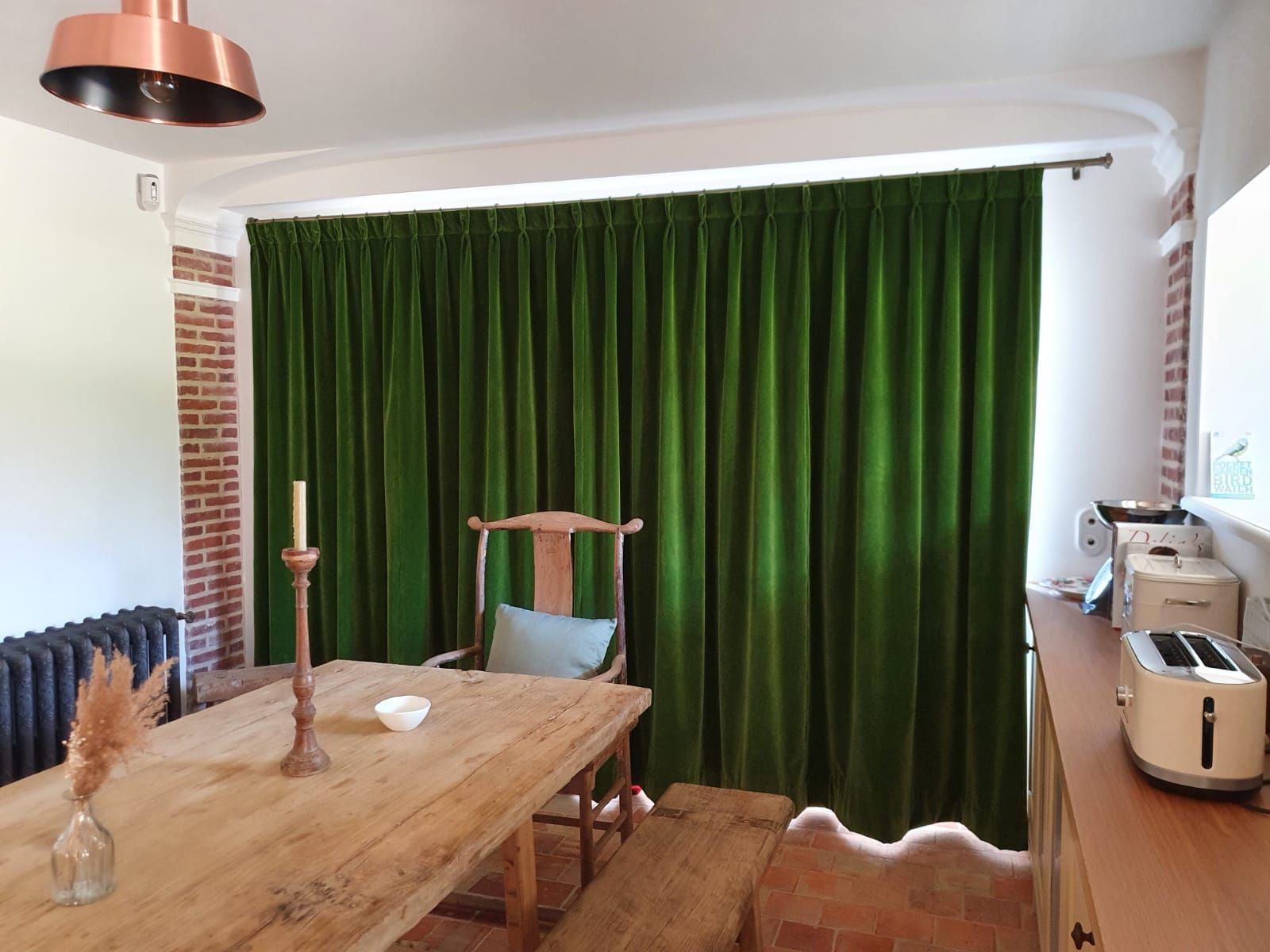 Des rideaux de velours vert drapent la salle à manger avec une table en bois, une chaise et un luminaire en cuivre. On y retrouve également des touches de brique et un comptoir avec des appareils électroménagers.