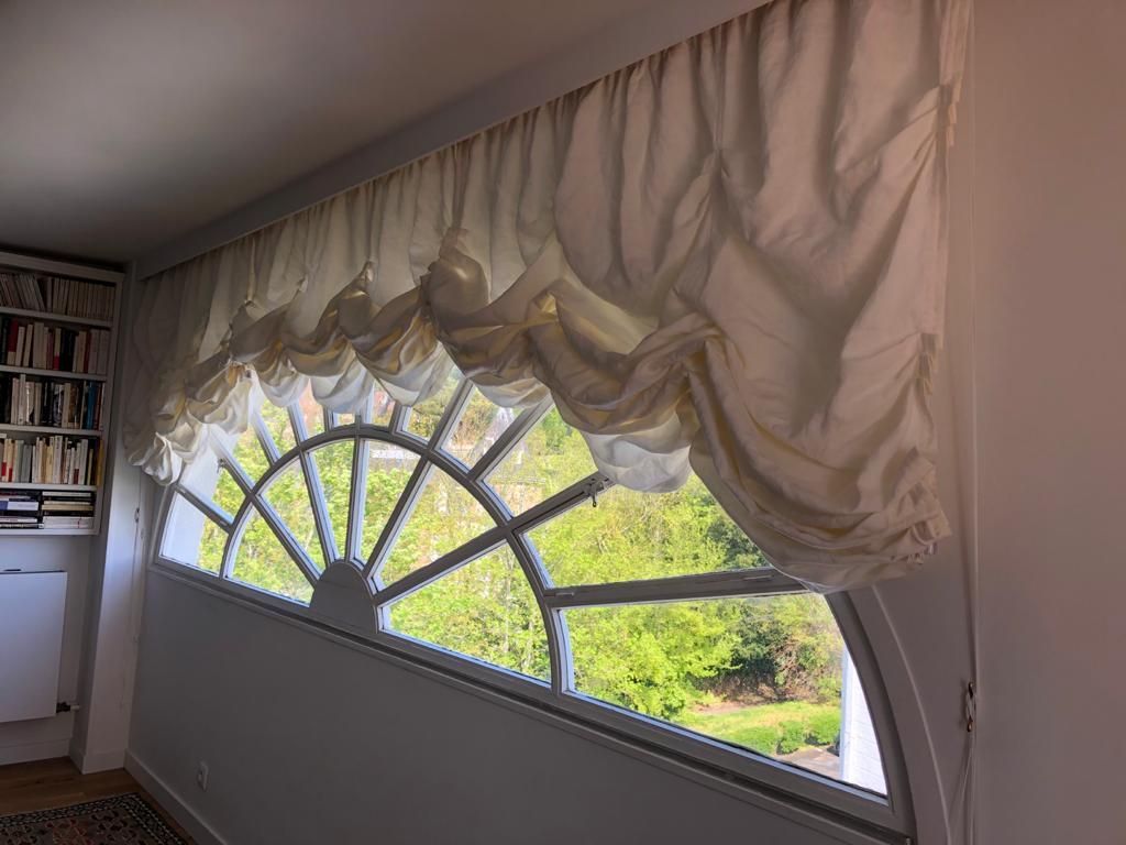 Un rideau blanc drapé sur une fenêtre en demi-cercle, avec vue sur la verdure à l'extérieur.