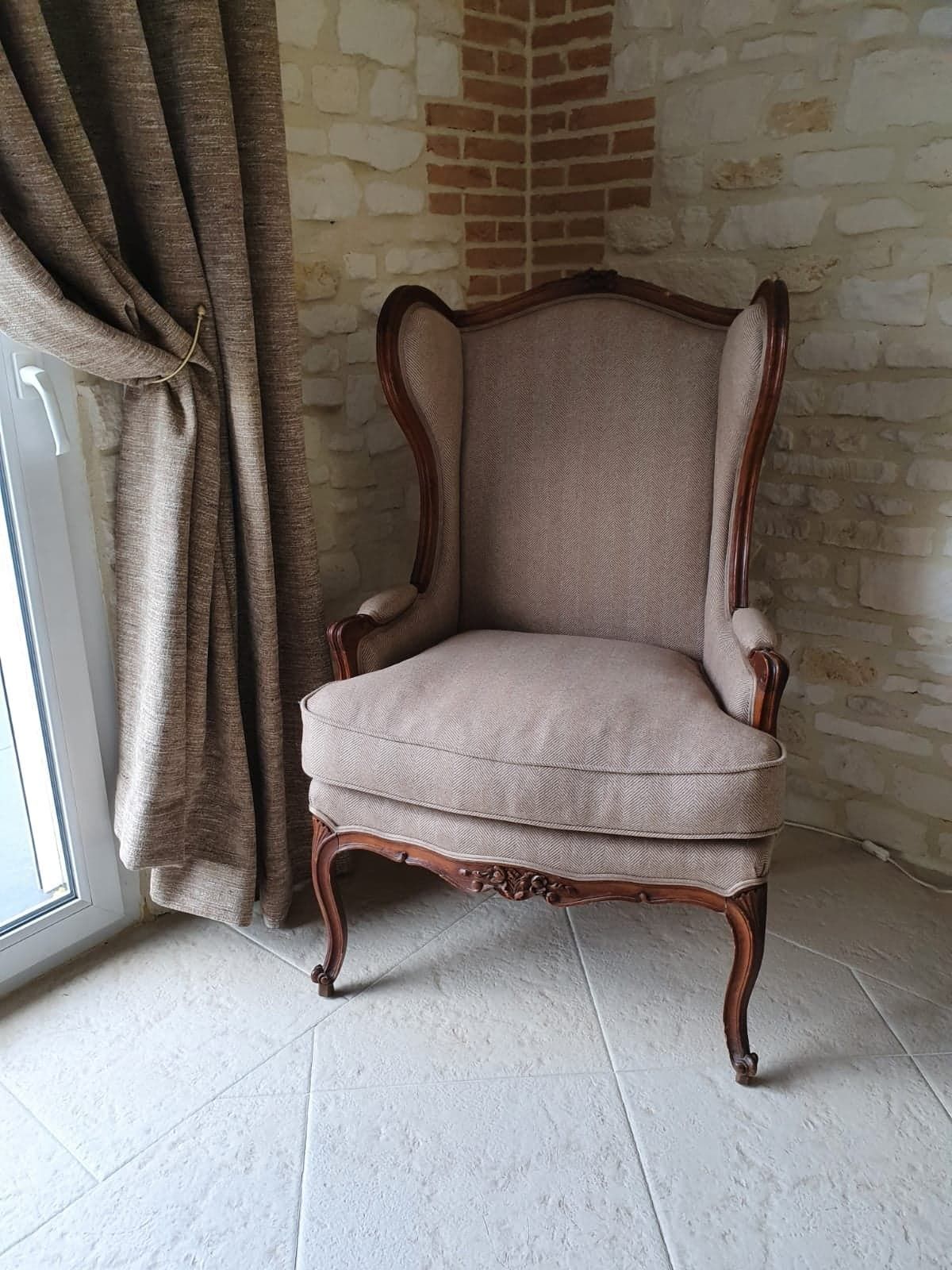 Une chaise à oreilles rembourrée avec des détails en bois se trouve près d'un rideau fermé et d'un coin en brique.