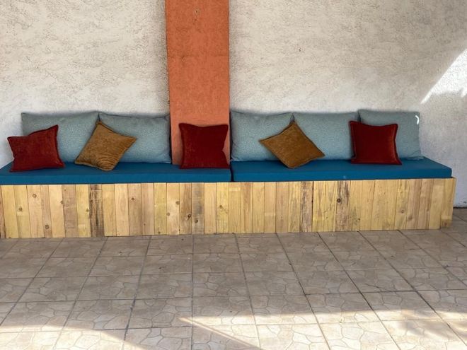 Un banc en bois avec des coussins bleus et des coussins colorés contre un mur clair. Le banc est situé à l'extérieur.