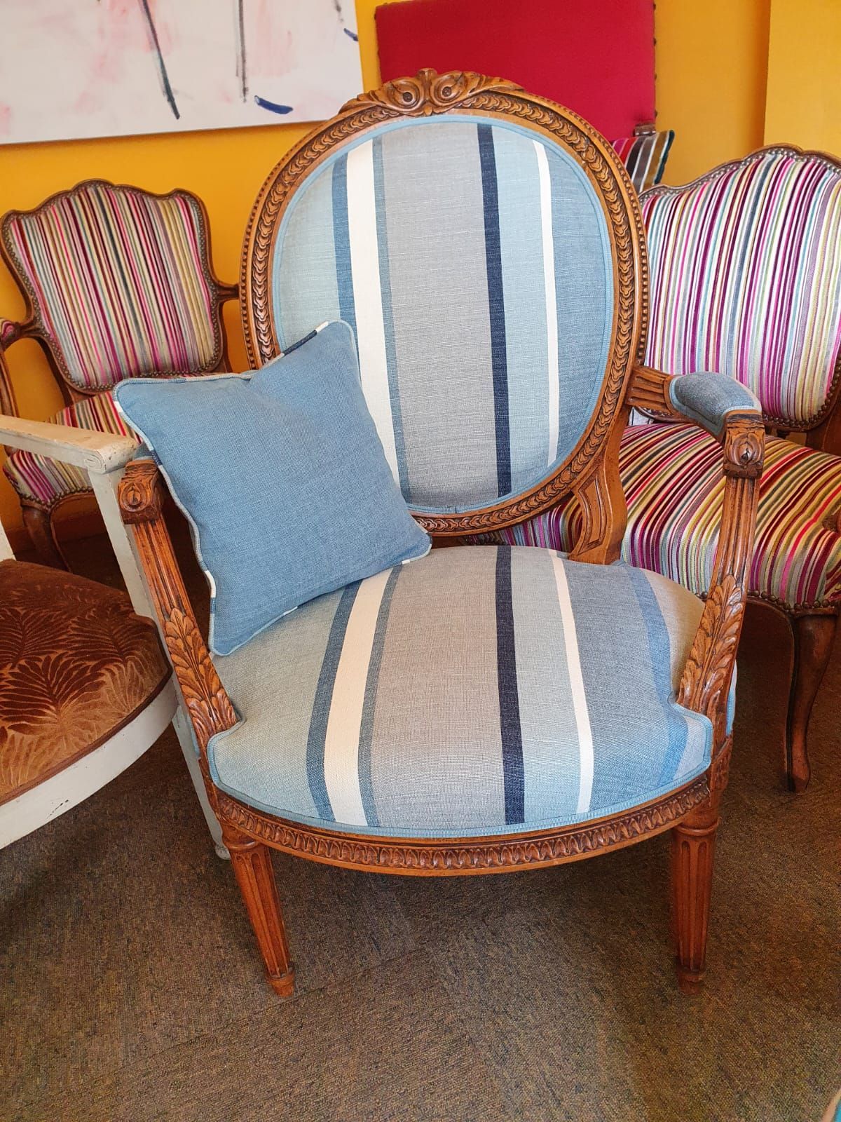 Une chaise en bois ornée, recouverte d'un revêtement rayé bleu et blanc, et un coussin assorti. D'autres chaises colorées sont visibles à l'arrière-plan.