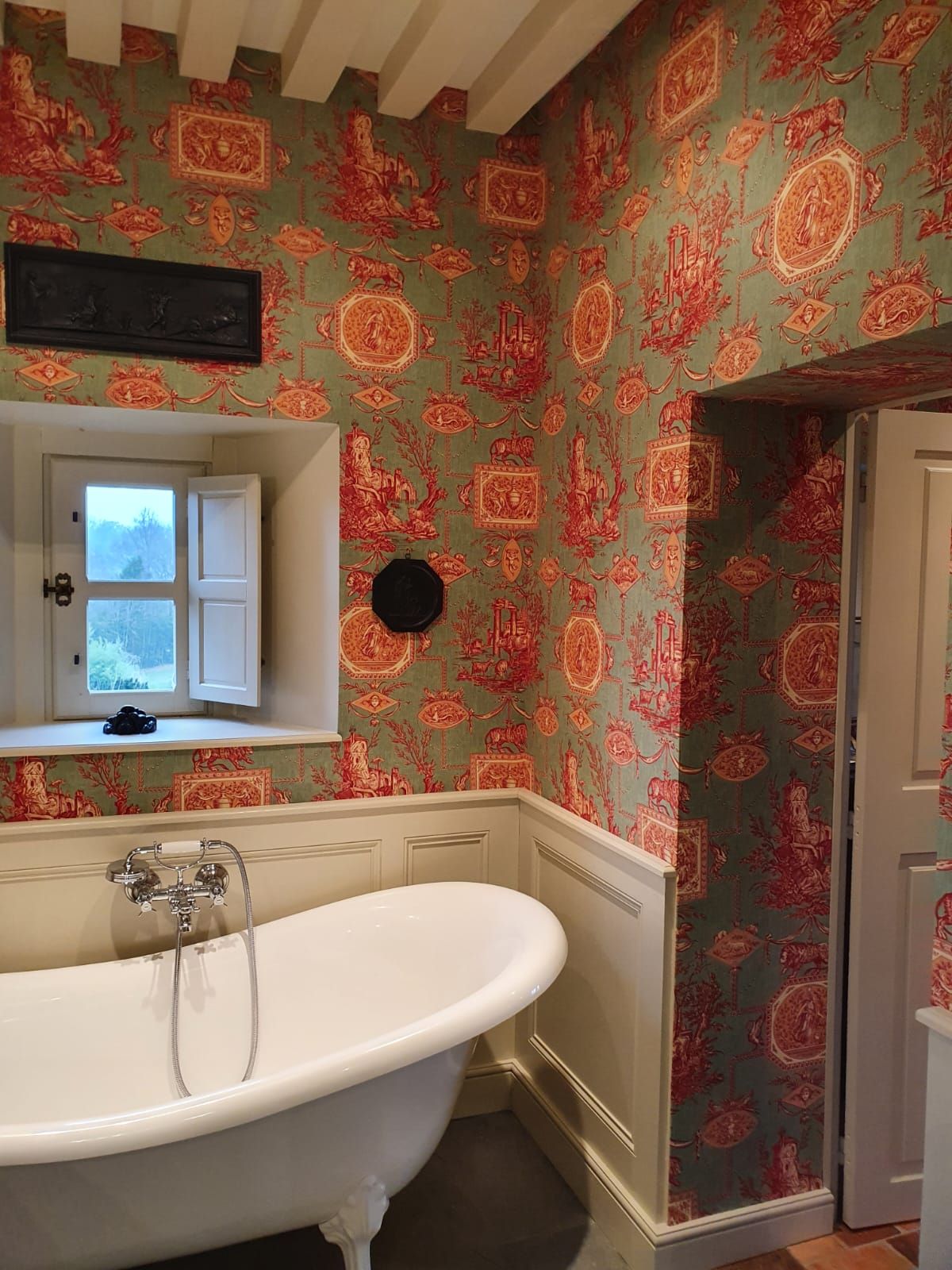 Salle de bain avec baignoire sur pieds, papier peint à motifs verts et rouges et une petite fenêtre.