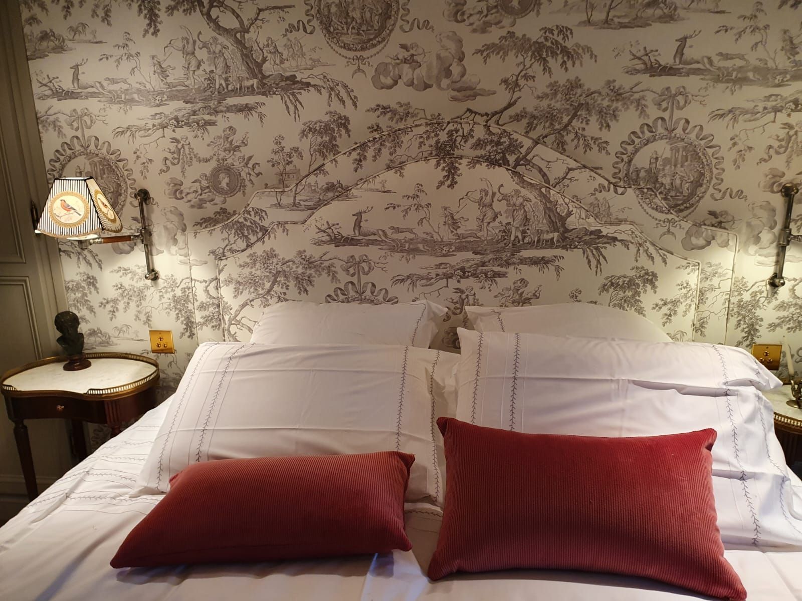Un lit aux coussins rouges trône devant un mur recouvert de papier peint en toile. Deux appliques et une petite table se trouvent de chaque côté.