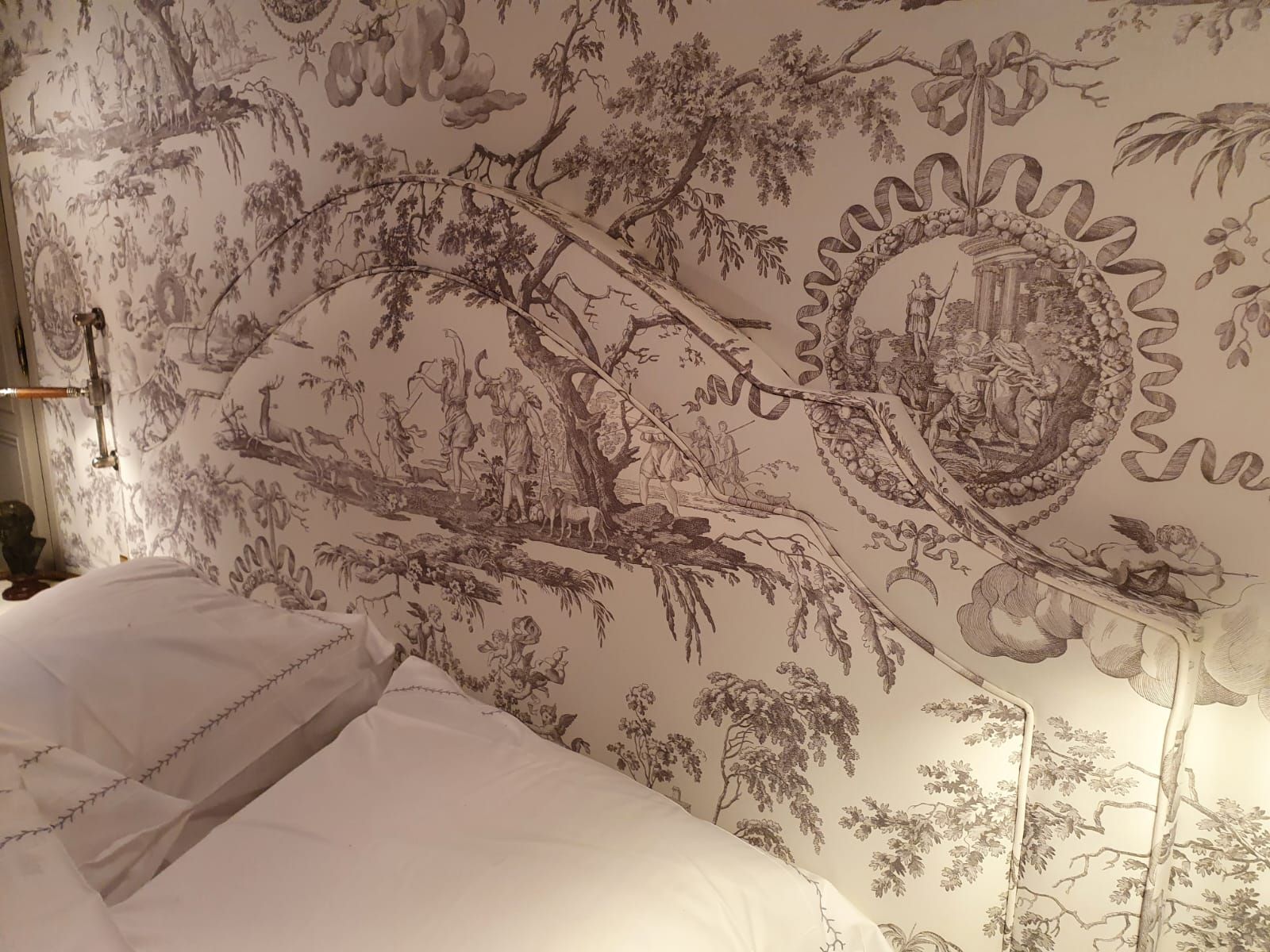 Chambre avec papier peint en toile de Jouy gris et blanc. Literie blanche visible.