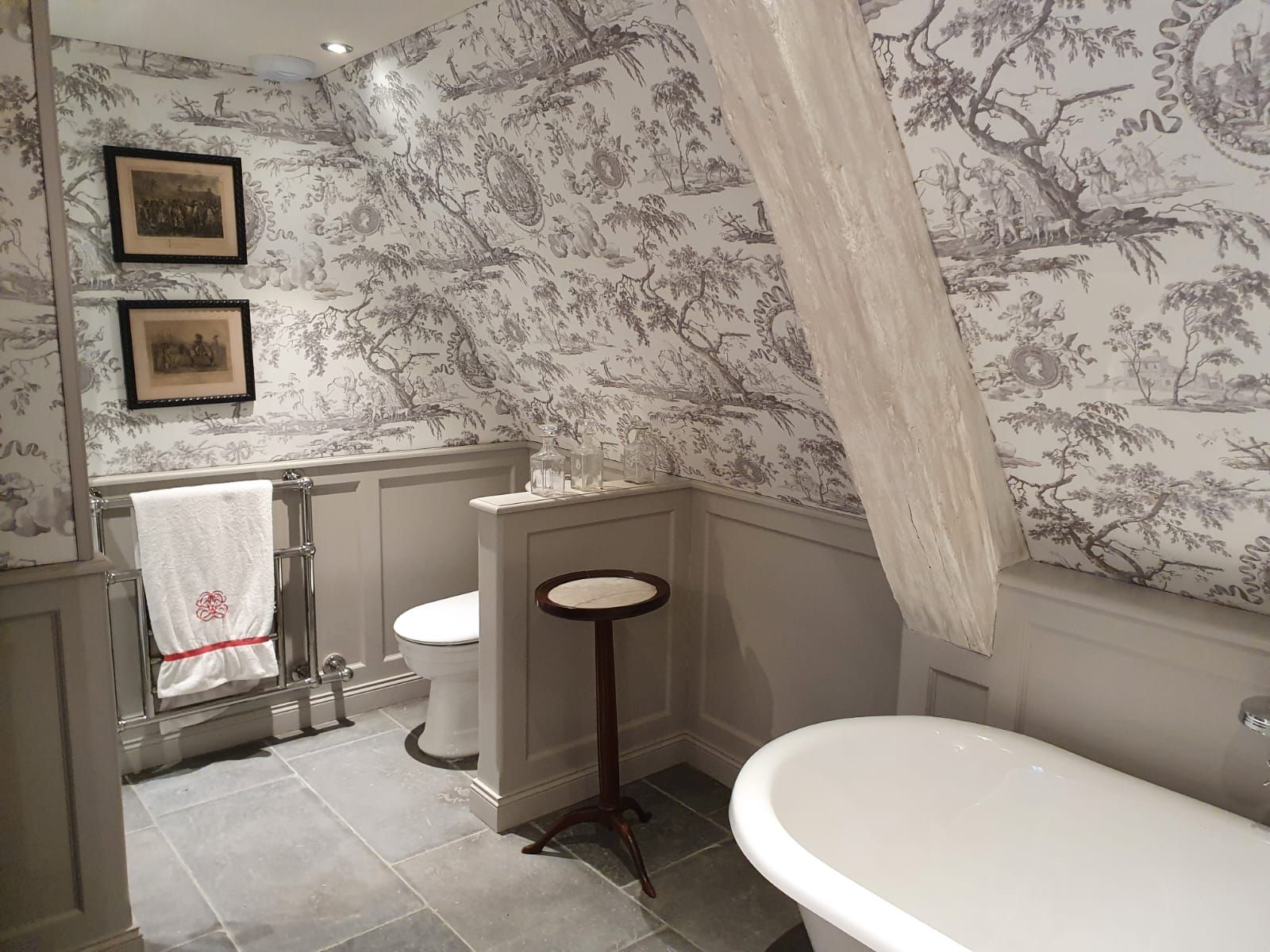 Une salle de bain avec des boiseries grises, des toilettes et une baignoire sur pieds. Les murs sont recouverts d'un papier peint en toile de coton noir et blanc aux allures pastorales.