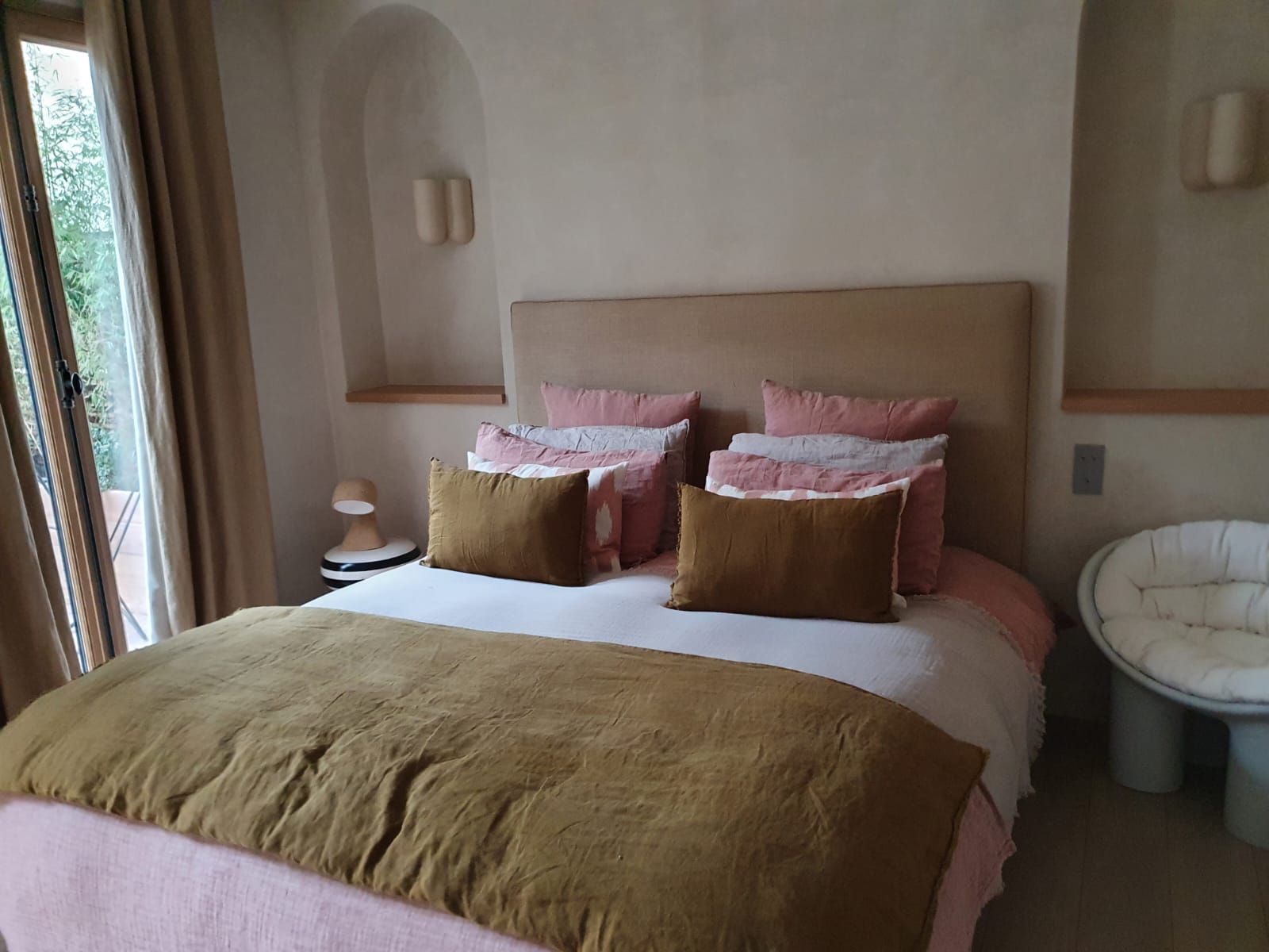 Une chambre avec un lit recouvert de draps roses et marron, des oreillers et une tête de lit. Il y a une fenêtre à gauche et une chaise à droite.