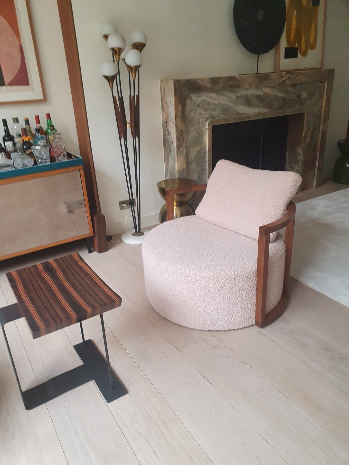 Un fauteuil rose, rond et bouclé se trouve dans une pièce avec une table d'appoint en bois, une cheminée et un chariot de bar.