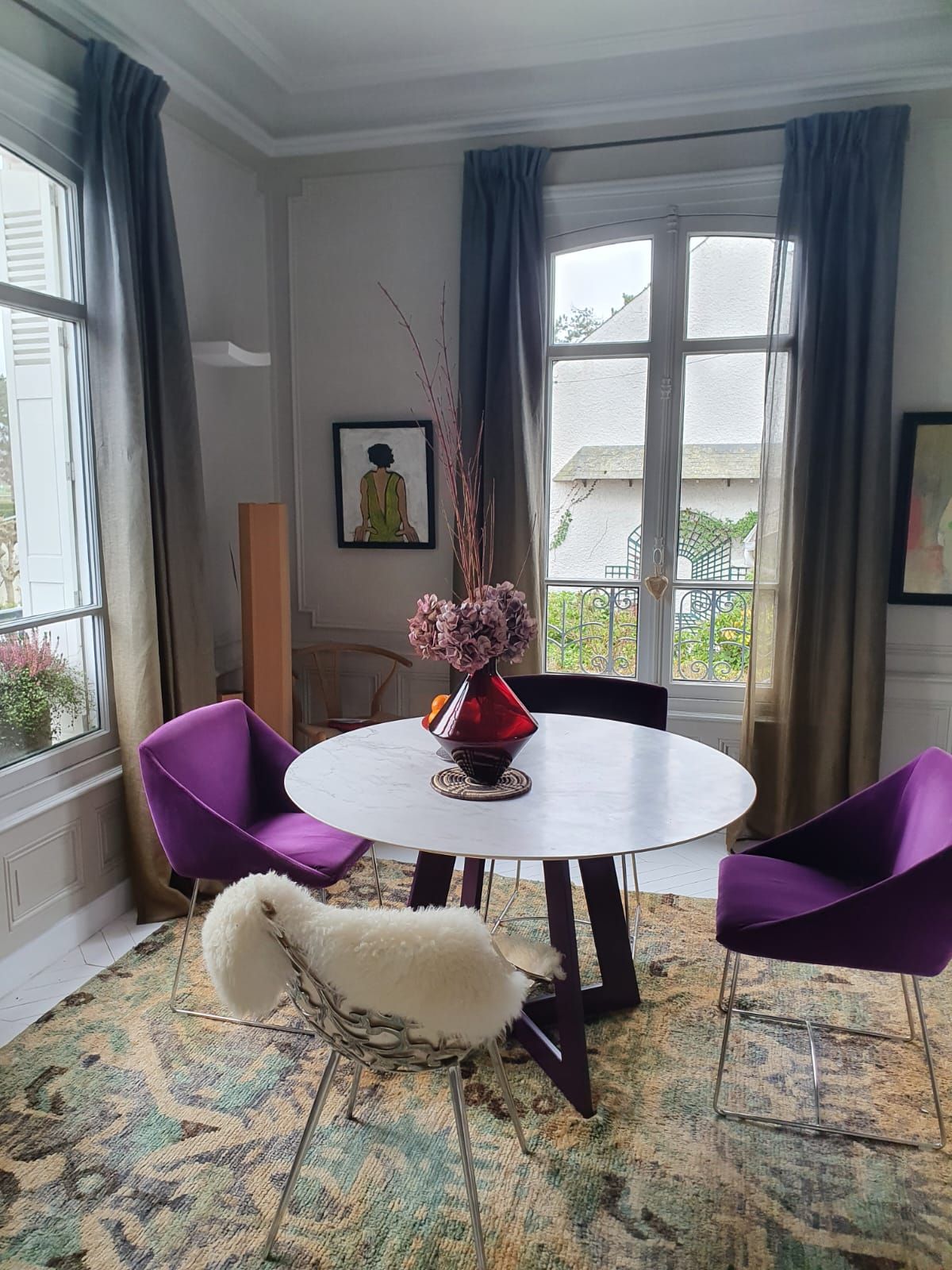 Une salle à manger avec une table ronde blanche, des chaises violettes et un tapis à motifs. Des fenêtres ornées de rideaux gris donnent sur un extérieur verdoyant.