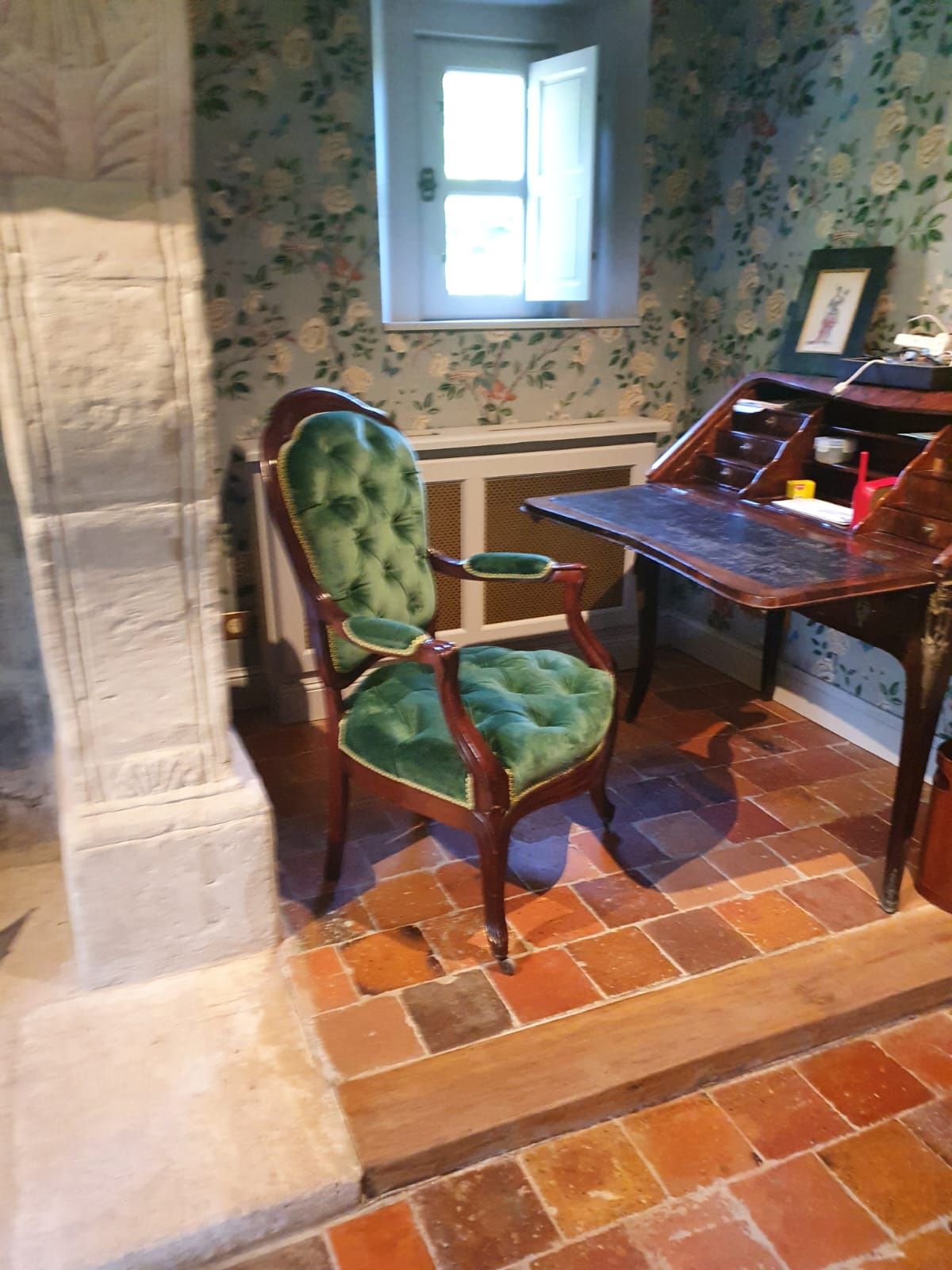 Une chaise en velours vert se trouve à côté d'un bureau en bois dans une pièce avec du papier peint à fleurs et une petite fenêtre.