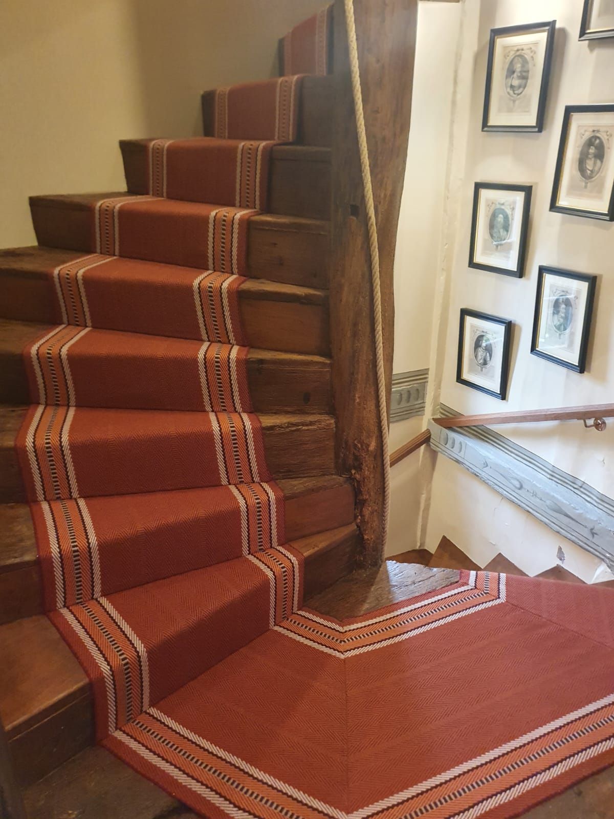 Escalier en bois avec tapis couleur rouille. Des portraits ornent les murs.