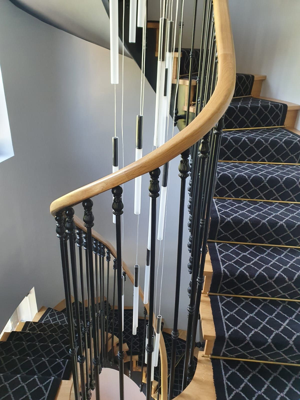 Un escalier en colimaçon avec moquette noire et rampe noire ornée. Une rampe en bois serpente le long des marches.