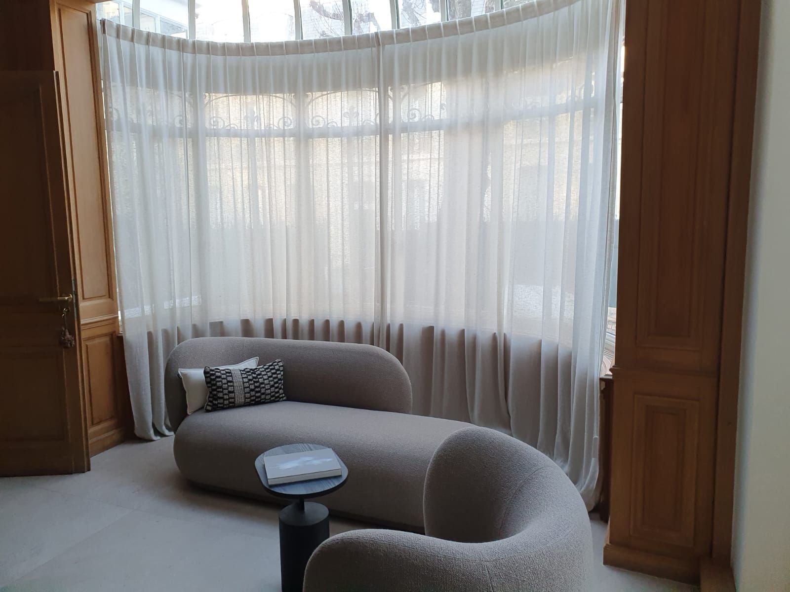 Un canapé incurvé de couleur claire trône devant une baie vitrée ornée de voilages. La pièce est agrémentée de boiseries et d'une petite table d'appoint.