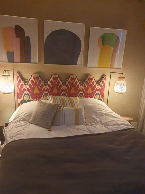 Chambre avec une tête de lit à motifs, des œuvres d'art au-dessus, deux lanternes suspendues et un lit avec des oreillers et une couverture marron foncé.