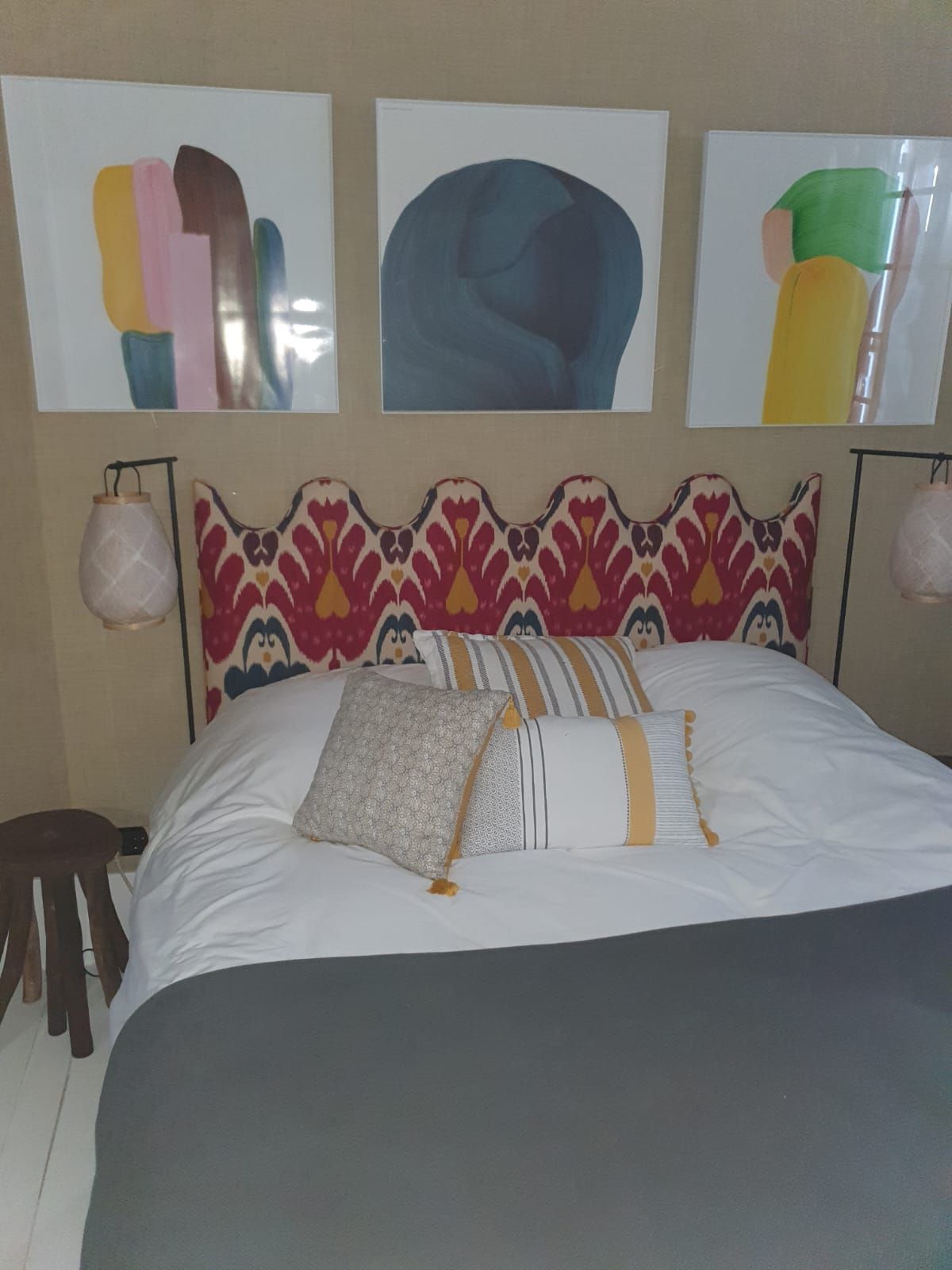 Chambre avec tête de lit à motifs et tableaux abstraits colorés au-dessus du lit. Couverture grise et oreillers aux motifs variés.