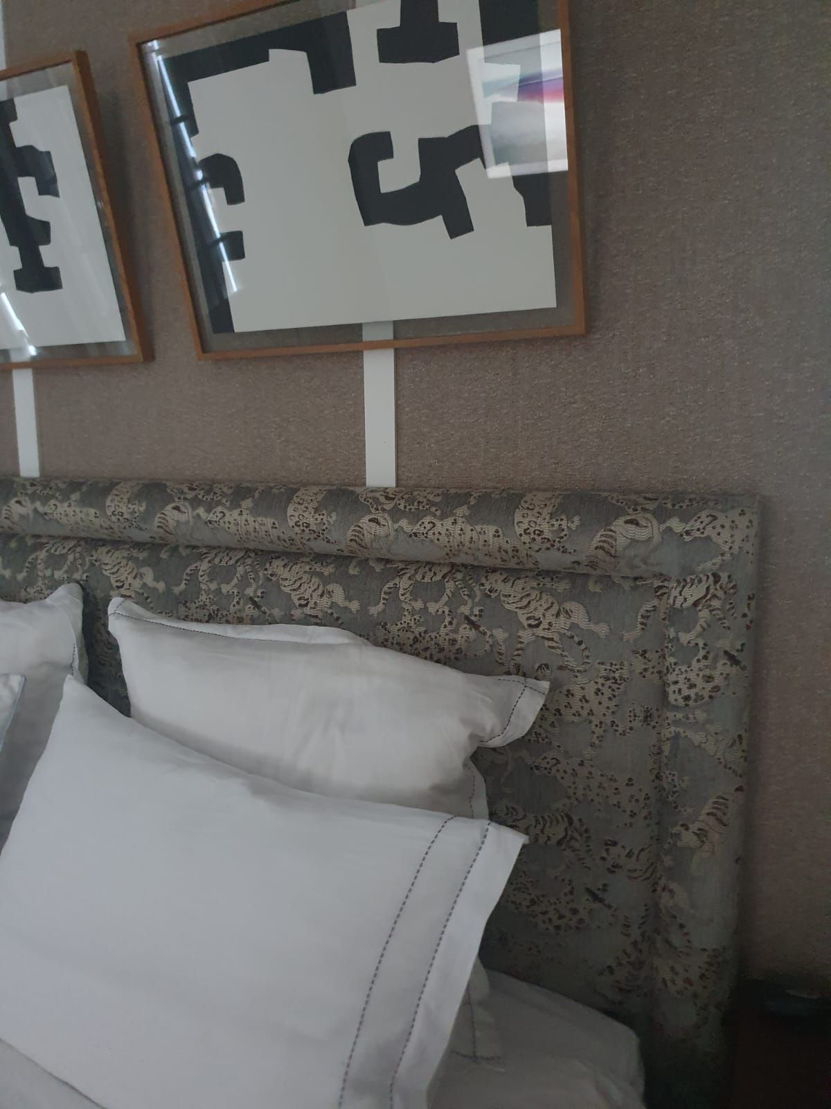 Tête de lit avec coussins blancs et tissu à motifs. Des œuvres d'art abstraites en noir et blanc sont accrochées au mur beige.