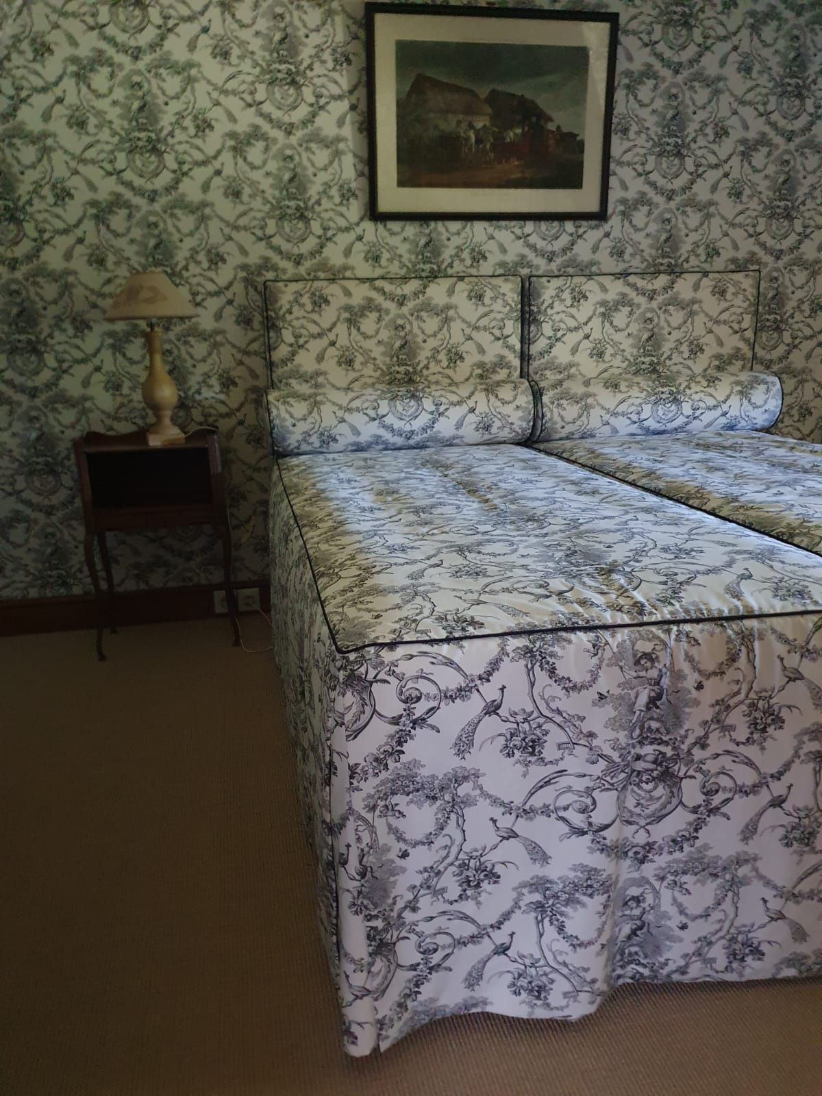 Chambre avec deux lits recouverts d'un motif floral, assorti au papier peint à motifs. Une petite table avec une lampe se trouve à proximité.