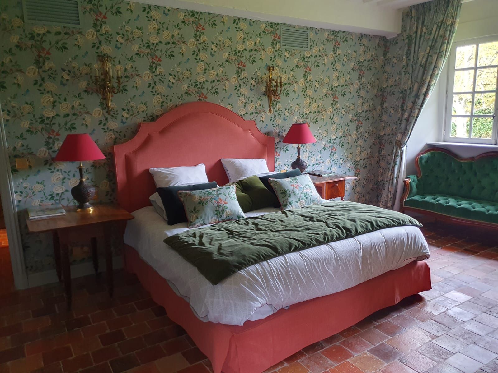 Une chambre avec un lit corail, du papier peint fleuri et un canapé vert. Des lampes rouges trônent sur les tables de chevet.