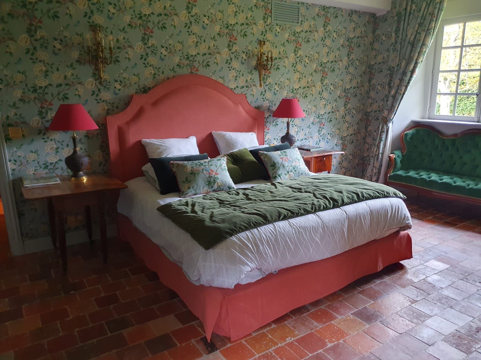 Une chambre avec un cadre de lit et une tête de lit corail, du papier peint floral, des lampes rouges et un canapé vert.