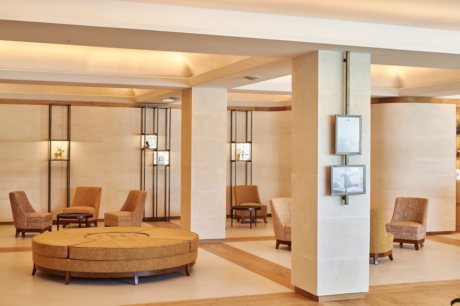 Un hall d'hôtel aux murs couleur crème, au mobilier beige et à l'éclairage encastré. Un grand pouf rond et plusieurs fauteuils sont disposés pour s'asseoir.