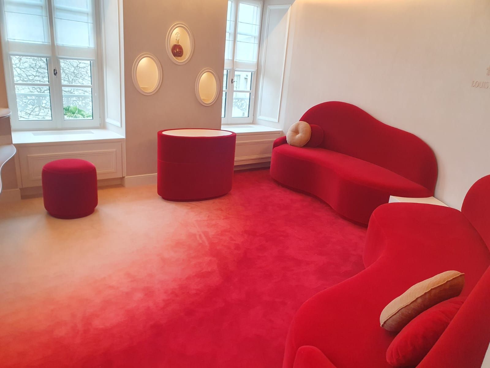 Sièges rembourrés rouges dans une pièce avec tapis rouge, deux fenêtres et un mur blanc avec un décor mural ovale.