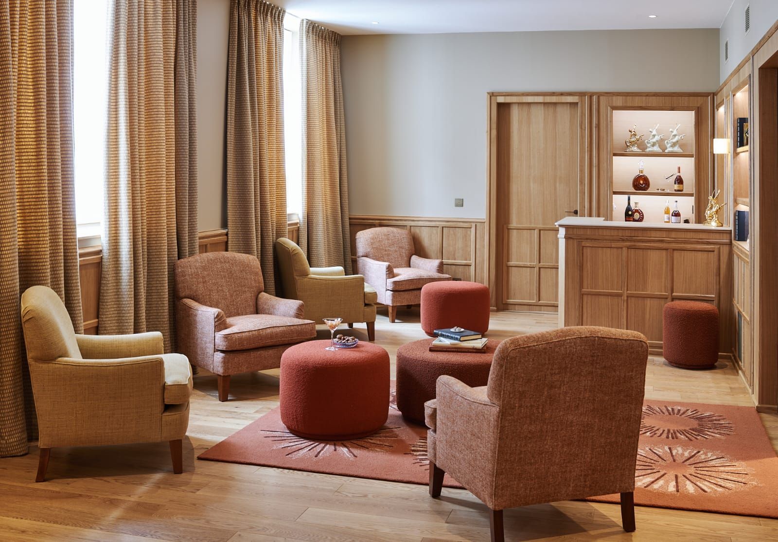 Un espace salon chaleureux avec plusieurs fauteuils rembourrés, des poufs et un petit bar. La palette de couleurs est chaleureuse, avec des nuances de pêche, de beige et de bois.