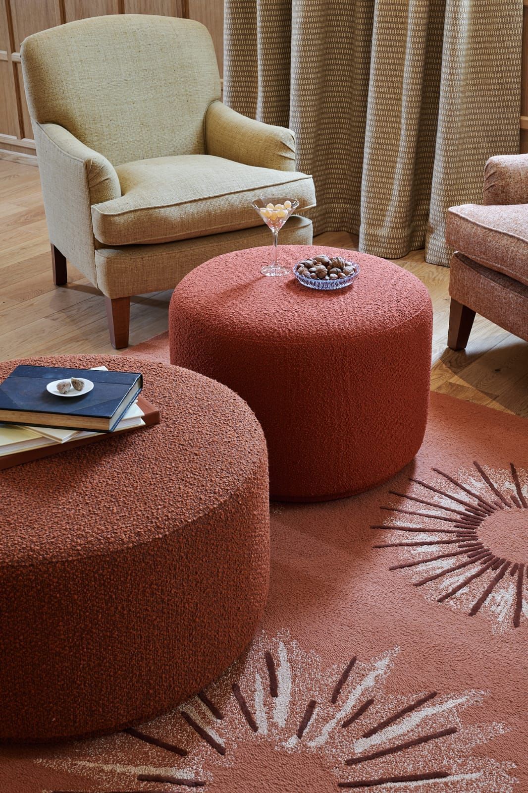 Deux poufs corail avec livres et en-cas dans une pièce avec tapis et fauteuils. On y retrouve également des rideaux neutres et un parquet en bois.