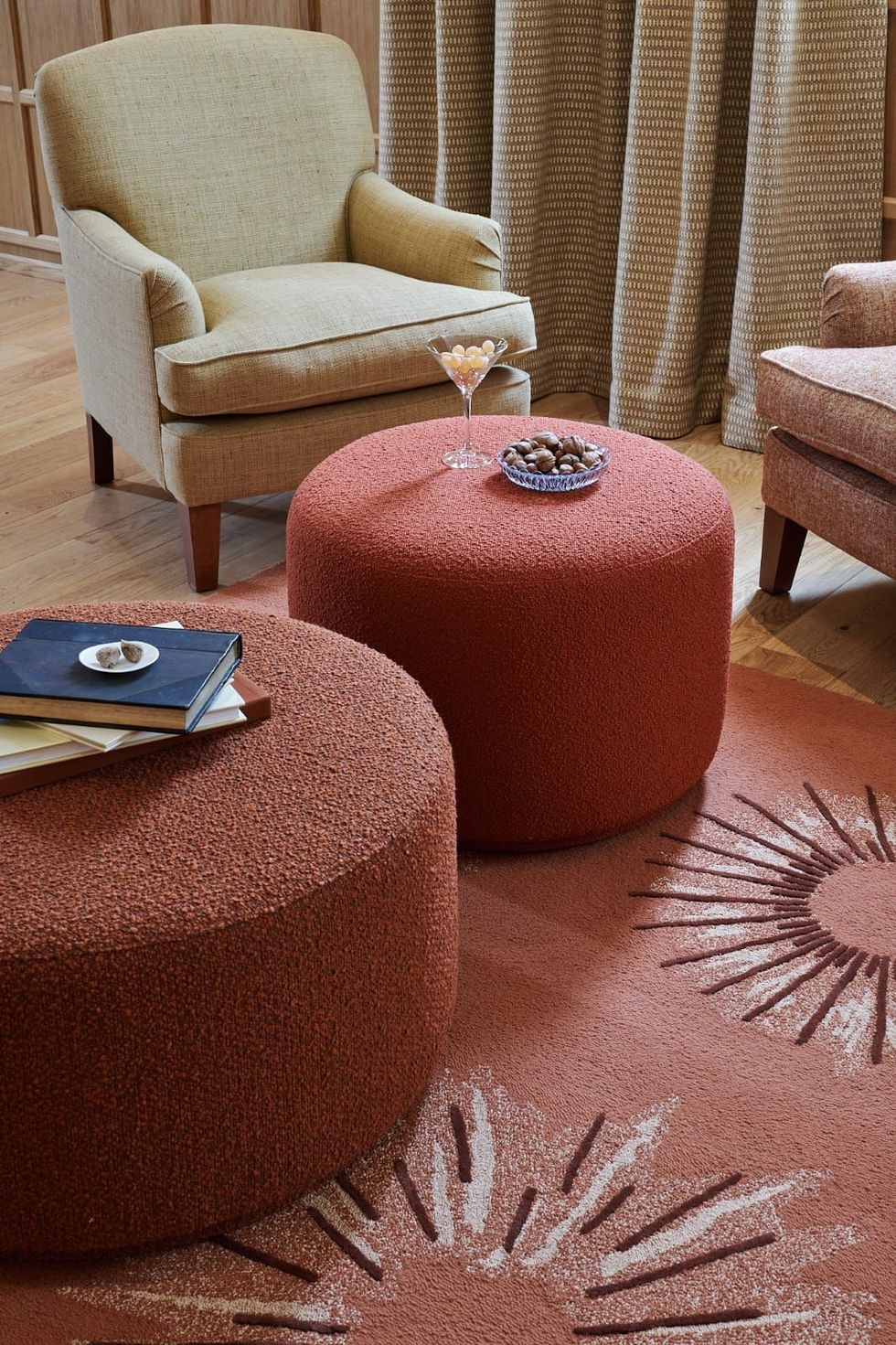 Deux poufs corail avec livres et en-cas dans une pièce avec tapis et fauteuils. On y retrouve également des rideaux neutres et un parquet en bois.