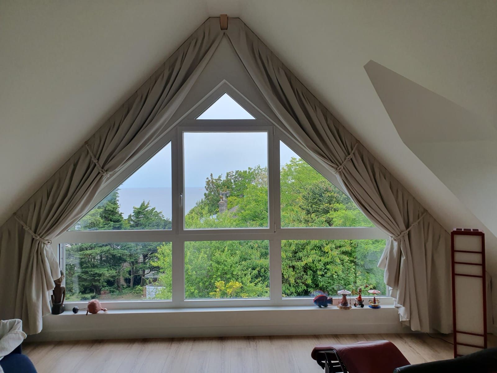 Une chambre mansardée avec une grande fenêtre triangulaire donnant sur des arbres verdoyants. Des rideaux beiges l'encadrent.