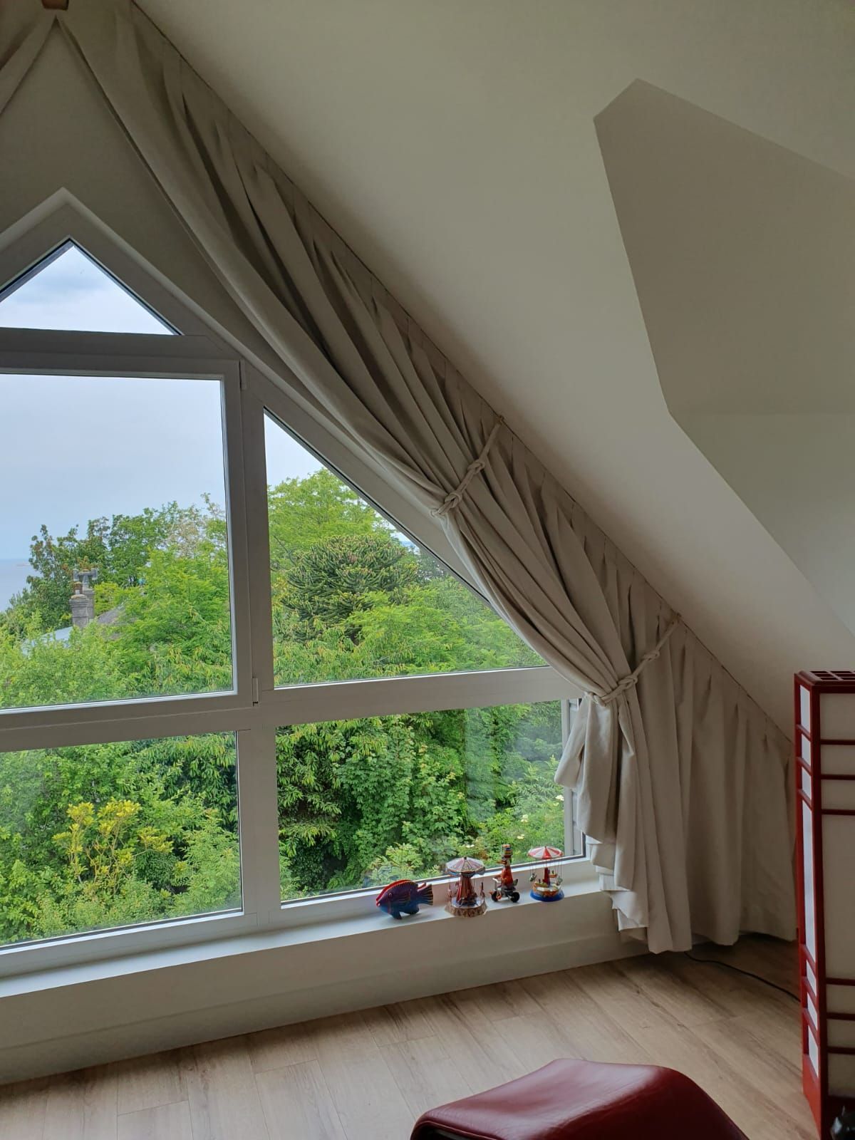 Fenêtre triangulaire avec rideaux beiges donnant sur des arbres verdoyants. On y aperçoit des murs clairs, un parquet imitation bois et une chaise rouge.