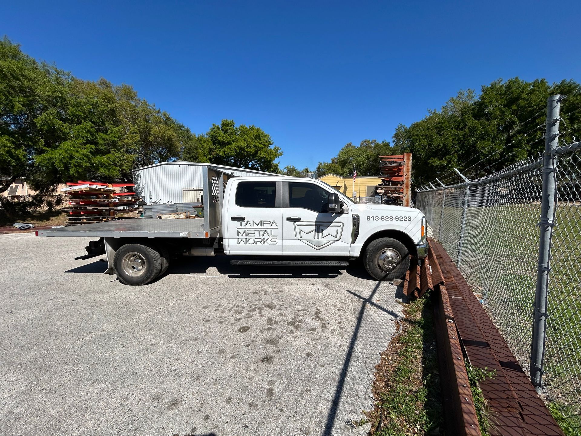 White flatbed truck parked in a gravel lot next to a chain-link fence on a sunny day.