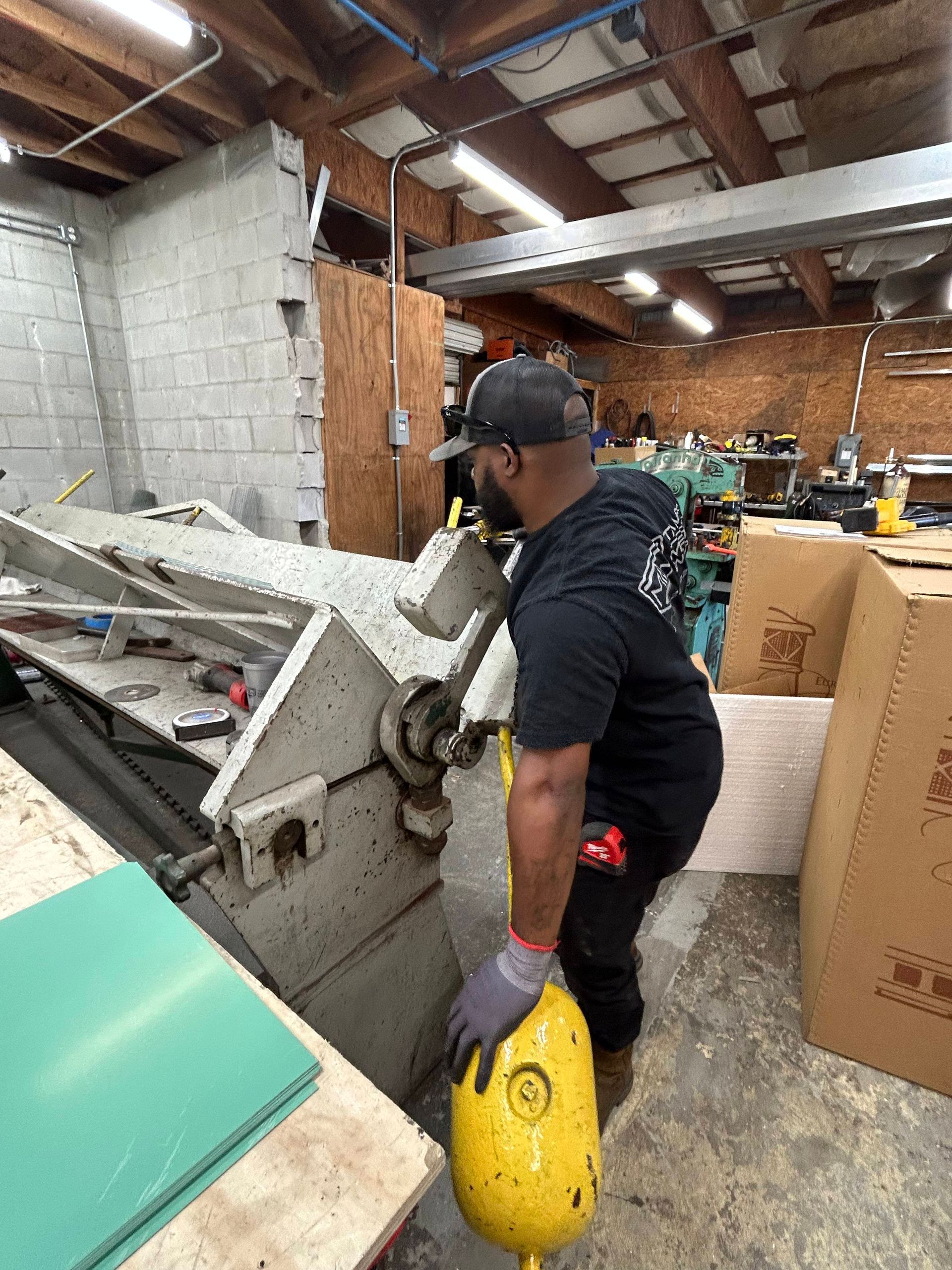 Man using a metal shear in a workshop; wearing a cap, lifting weight.