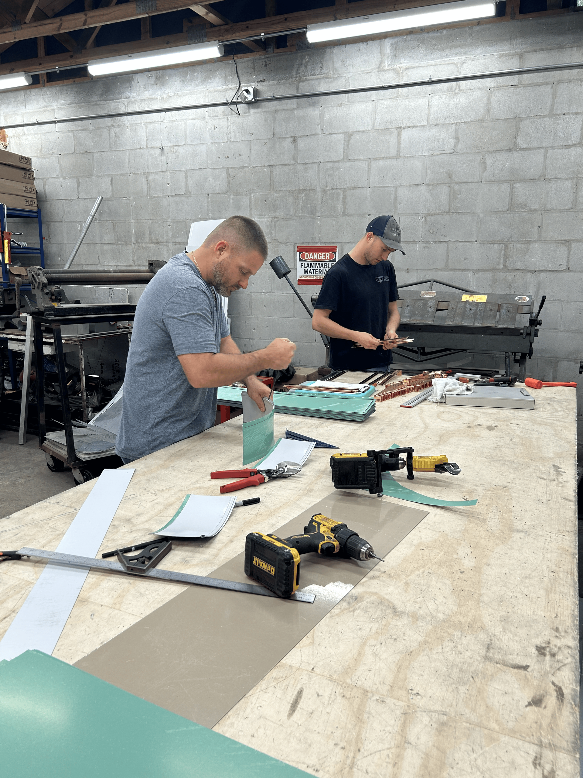 Two men work at a workbench, cutting metal sheets in a workshop.