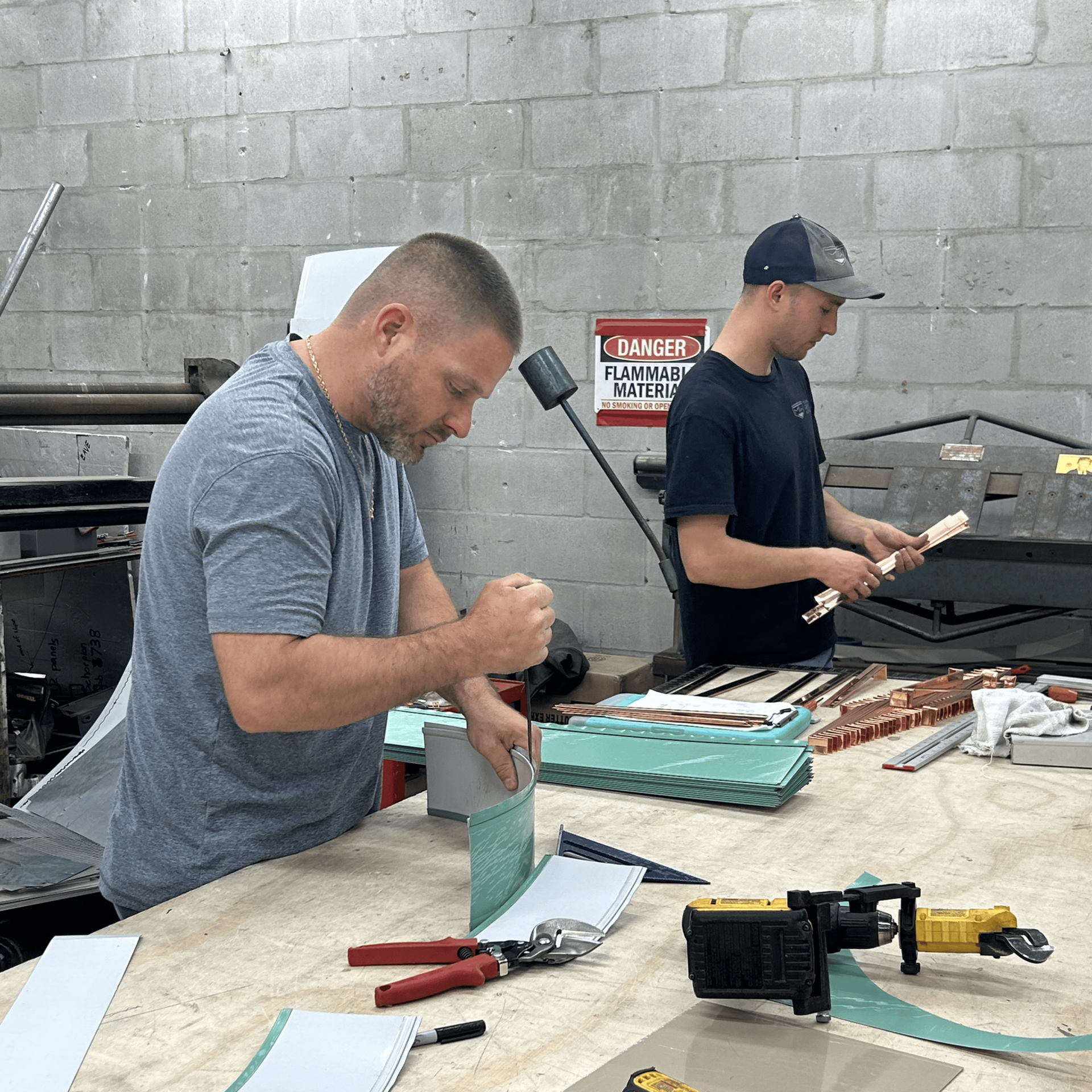 Two men work in a workshop. One cuts metal with shears, the other inspects copper pieces.