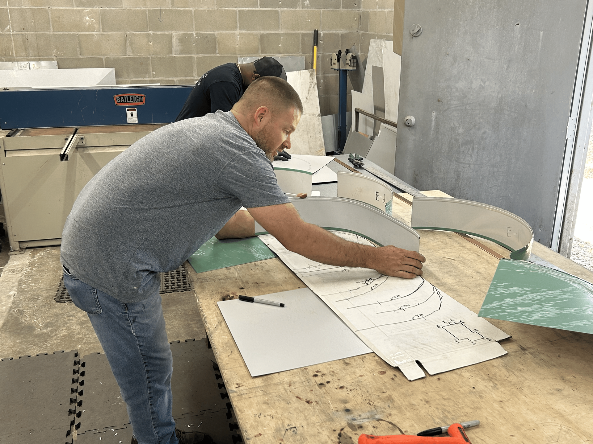 Man tracing curved shape on template, preparing to cut metal in workshop.