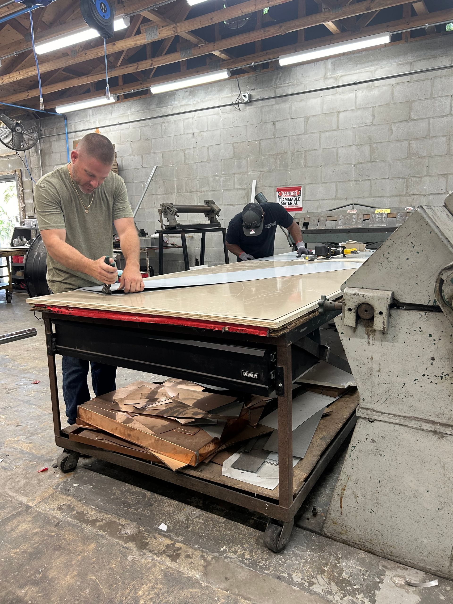 Two men working with metal sheets in a workshop; one cuts while the other bends over.