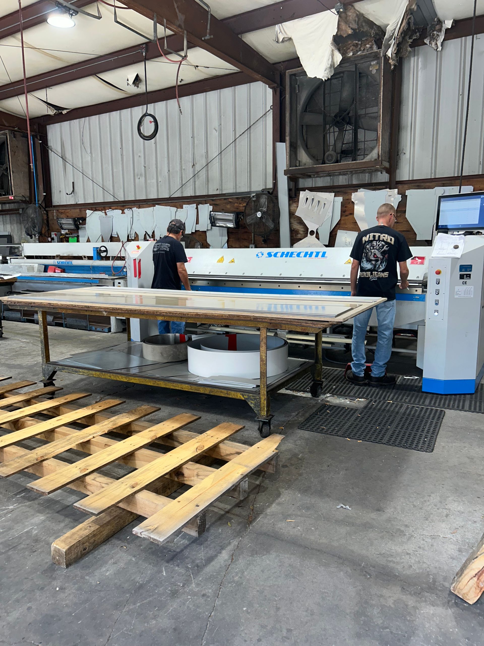 Two men operate a large industrial machine in a workshop. A wooden pallet sits in the foreground.