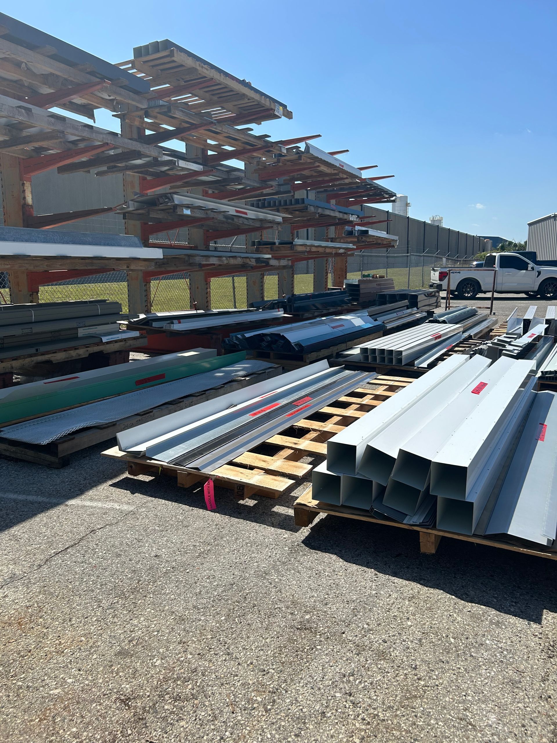 Metal building materials on pallets, racks, and a gravel lot. Blue sky, sunny day.