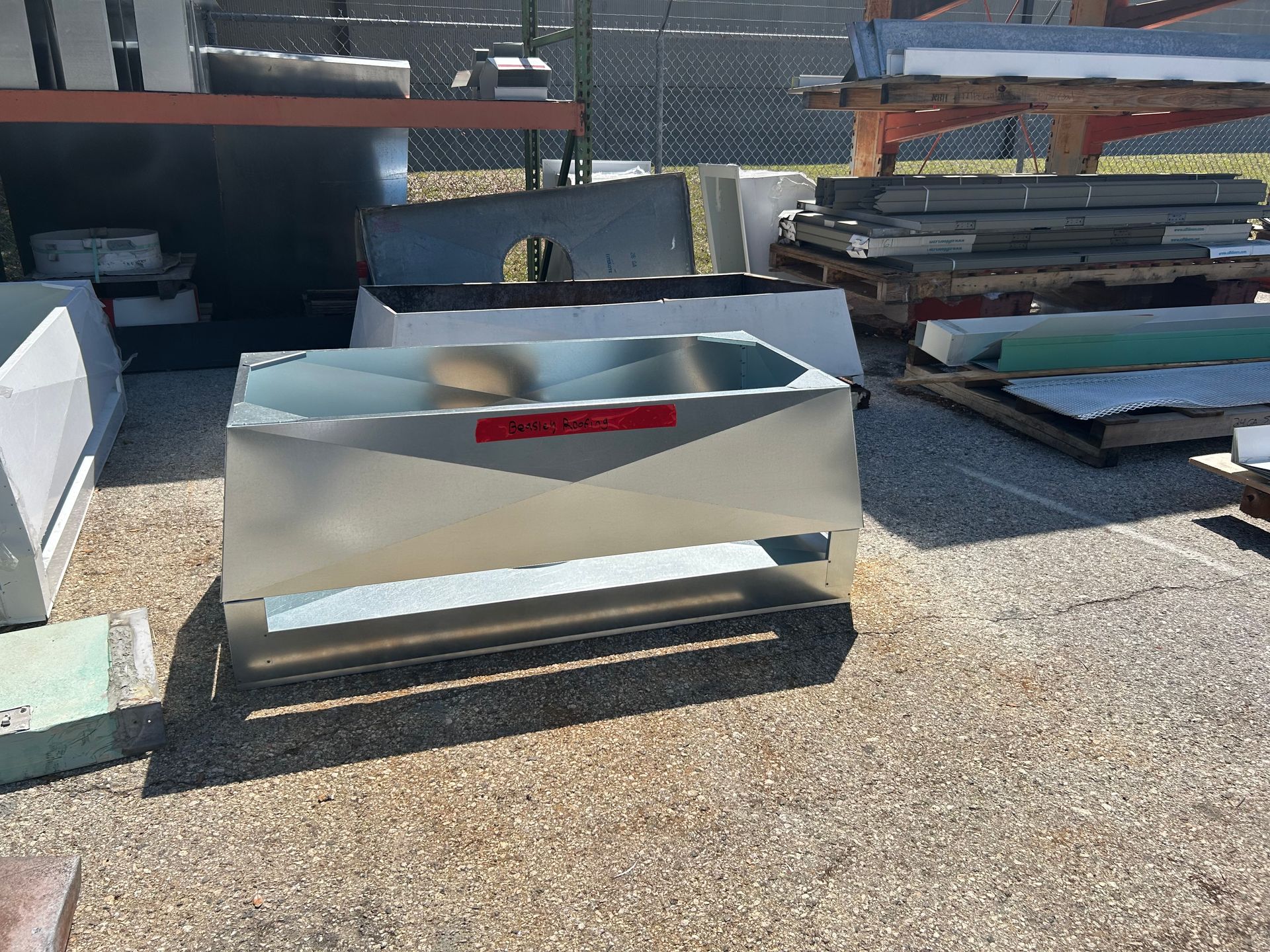 A stainless steel rectangular bin with a red sticker sits on gravel outside with other metal pieces.