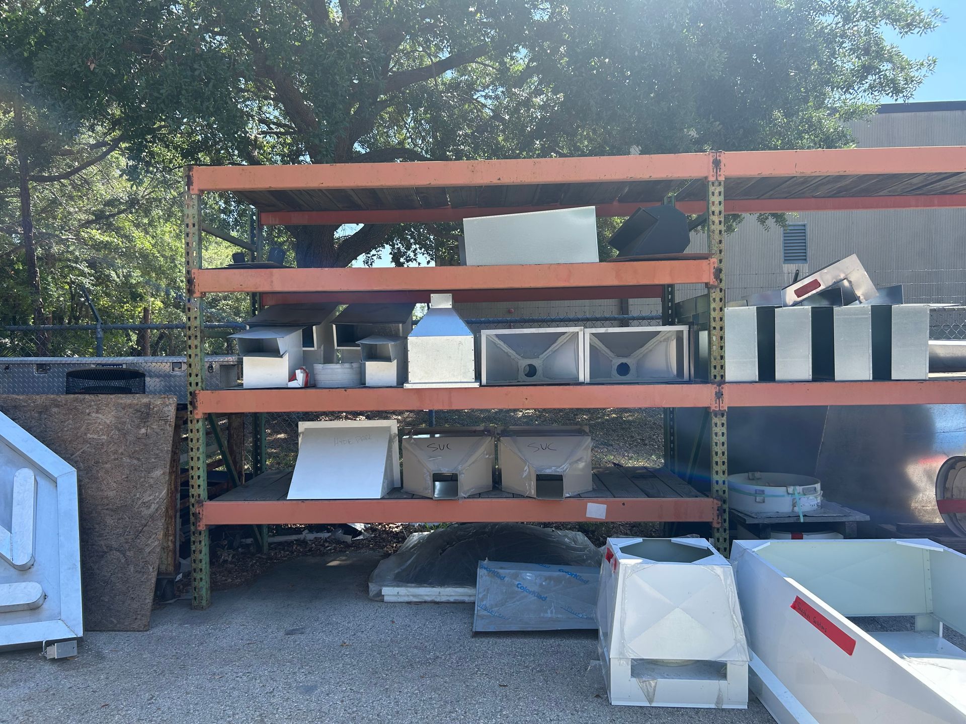 Shelves holding various white and gray industrial parts outside in a sunny area, near trees and a building.