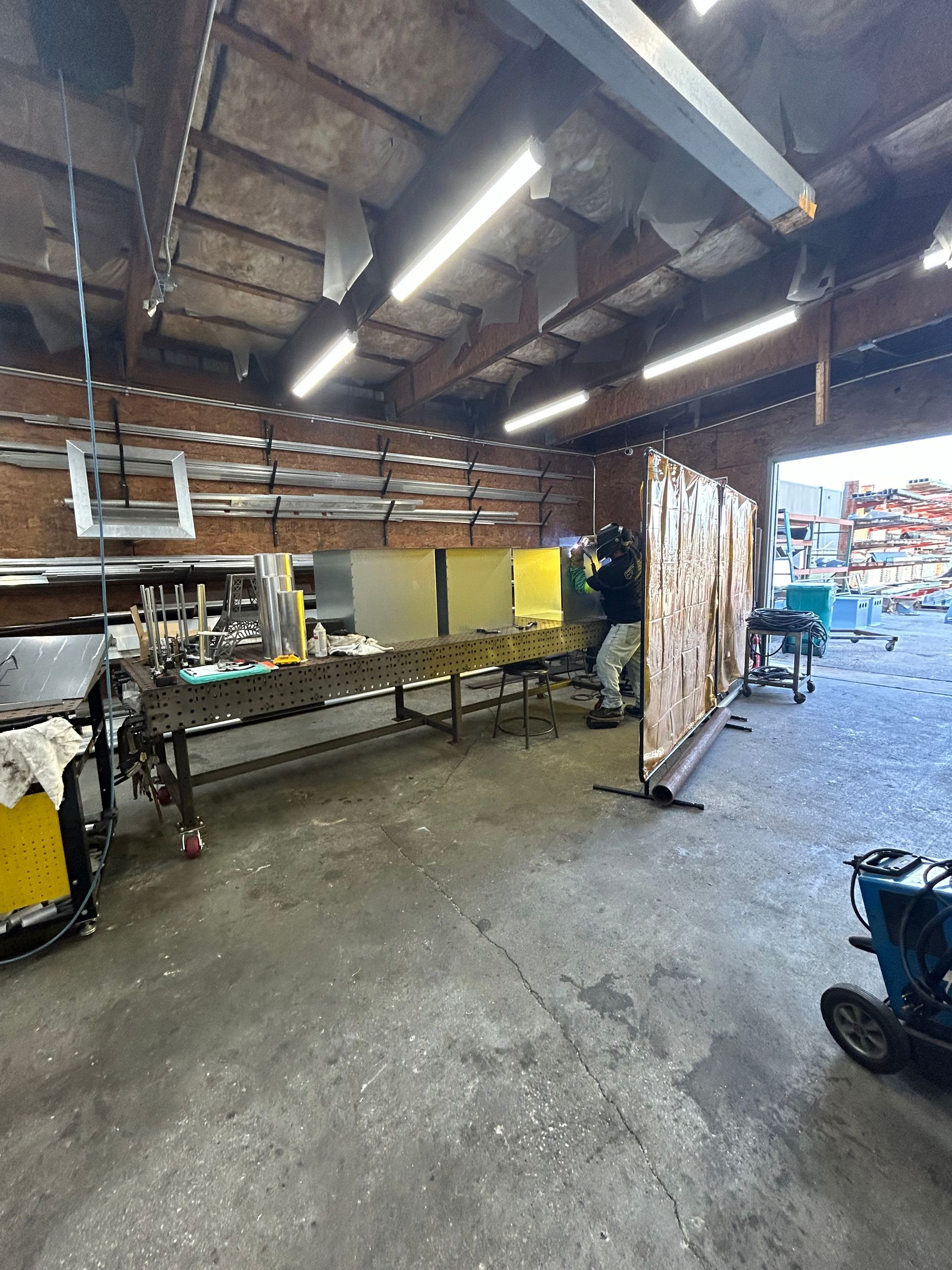 A person welding in a workshop, behind a protective screen. Metal tables and tools are visible.