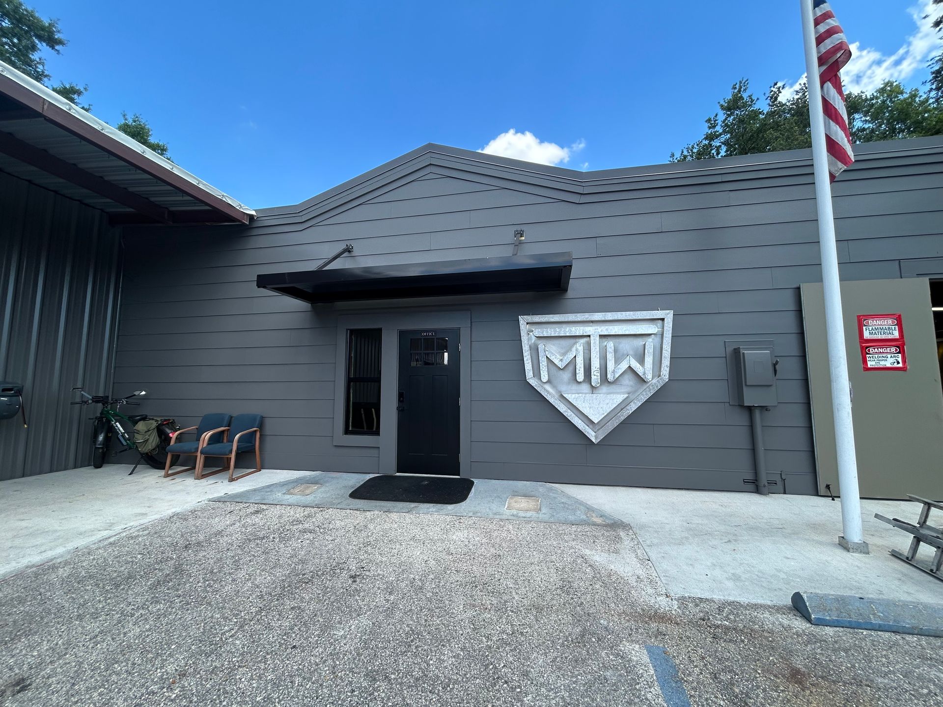 Gray building with a shield logo, an American flag, and a gravel driveway under a blue sky.