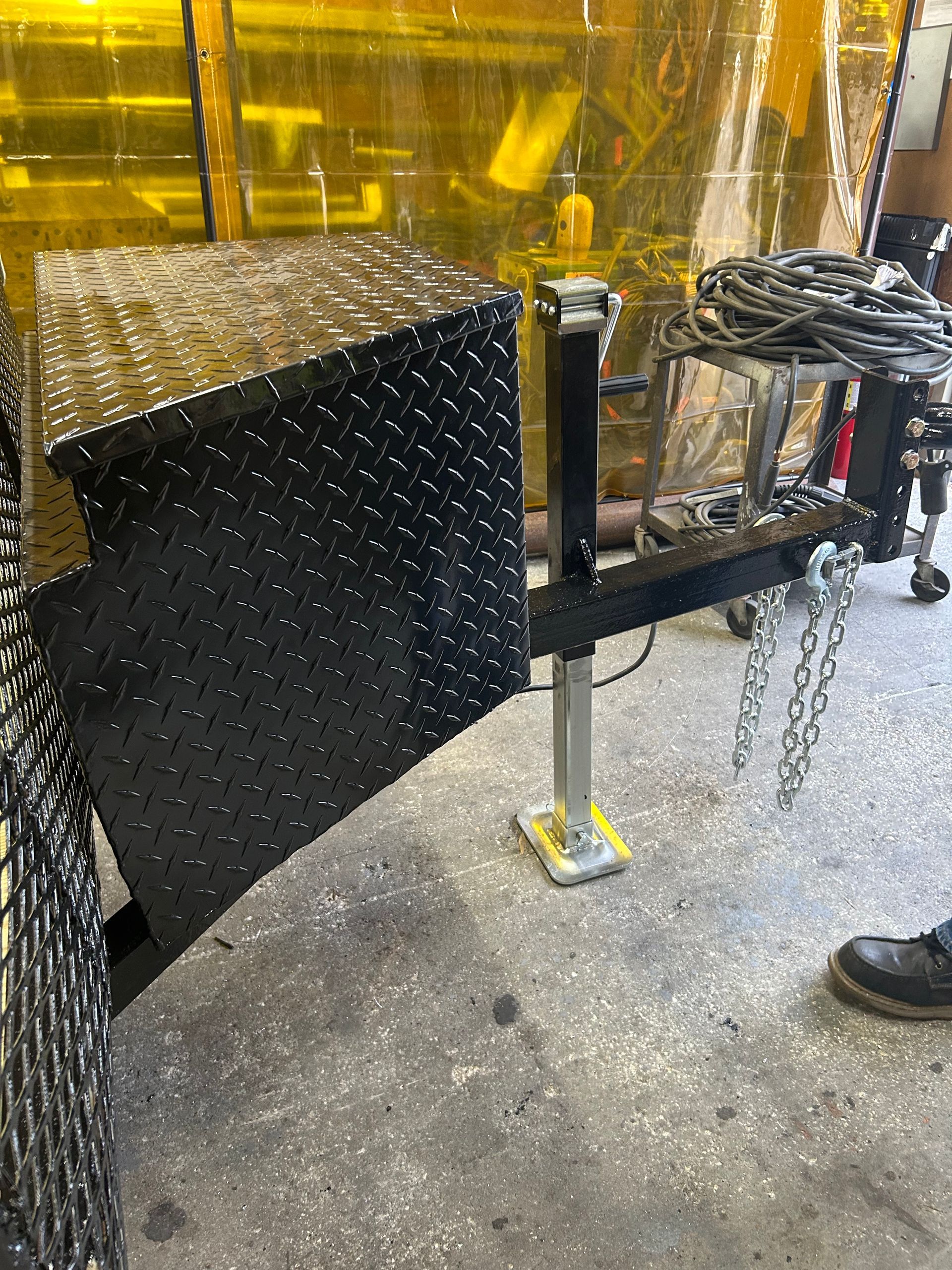 Black metal trailer with a toolbox and jack on a concrete floor.