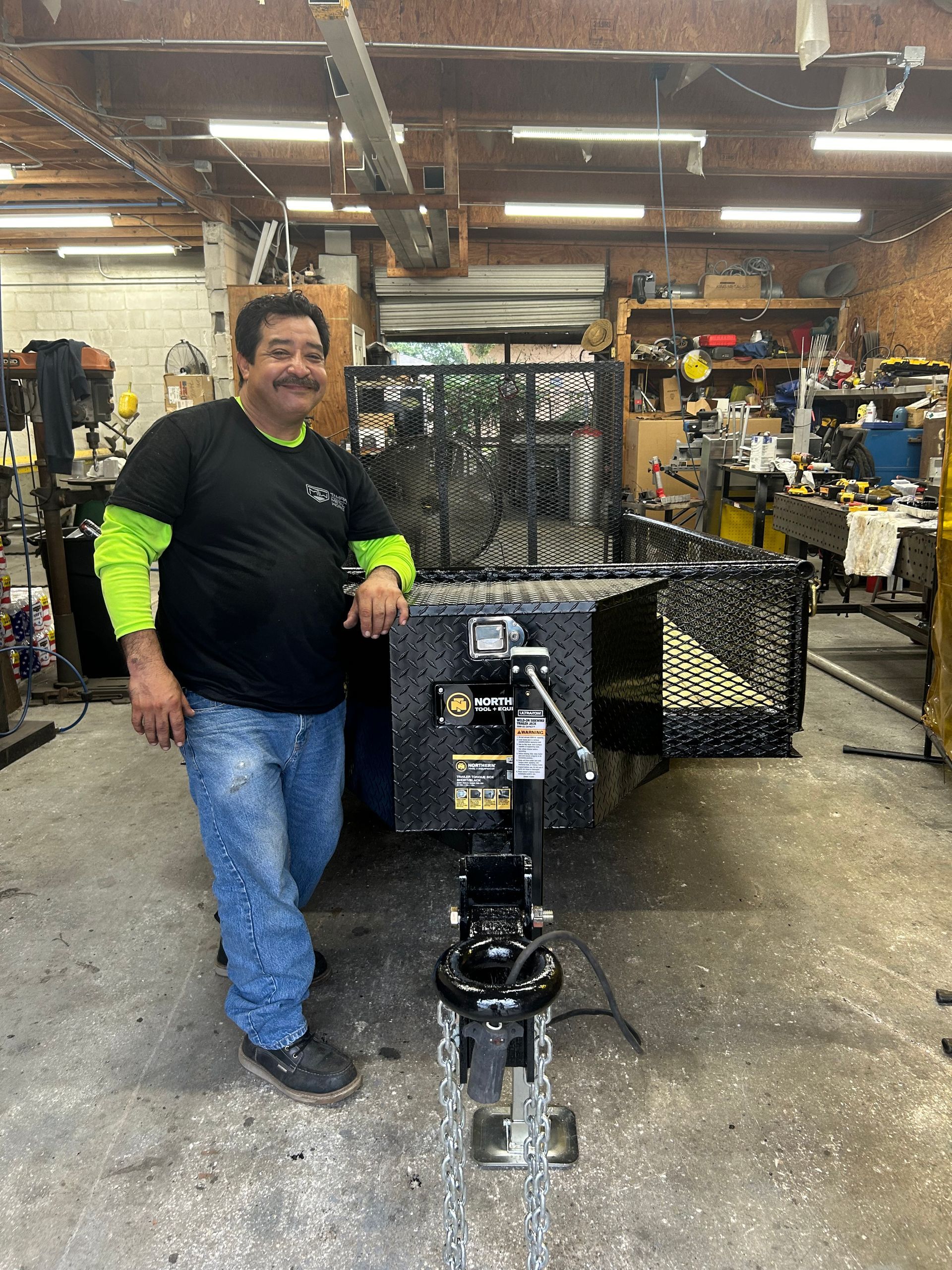 Man in shop next to trailer; he leans on it, wearing a black shirt and jeans. Industrial setting.