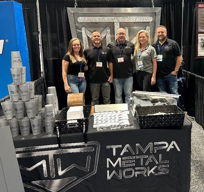 Tampa Metal Works team at a booth, smiling, with cups and products on the table, metallic logo.