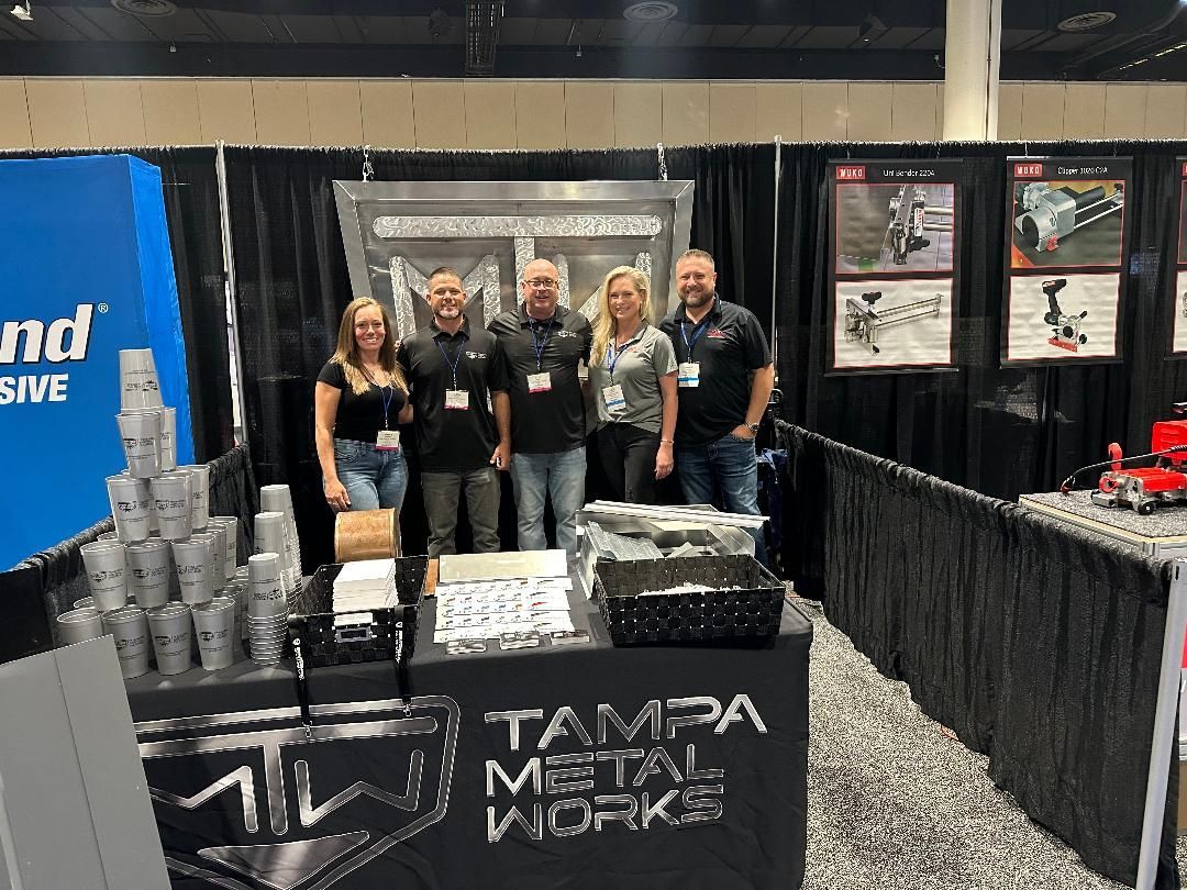 Group of five at the Tampa Metal Works booth, smiling, behind a table with products at a trade show.